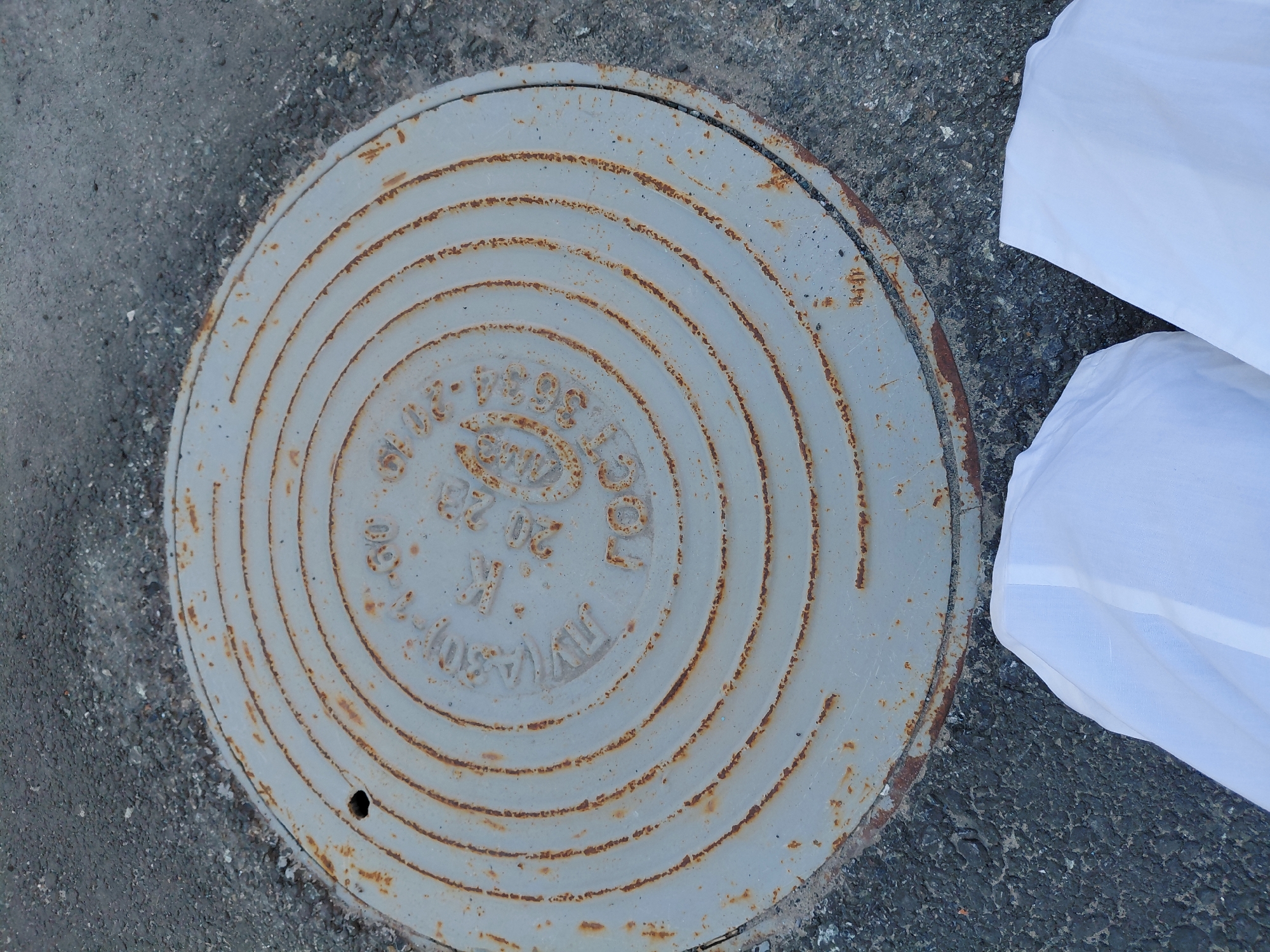 A slightly tilted, high-angle shot captures a circular, light grey metal manhole cover embedded in dark grey asphalt and gravel. The manhole cover is heavily worn, showing extensive reddish-brown rust along its concentric raised rings and overall surface, indicating oxidation. A small, dark circular hole is visible near the bottom-left edge of the cover.
On the right side of the frame, two white, folded objects, possibly fabric sheets or plastic bags, are partially visible, lying on the asphalt next to the manhole cover. The scene is static, illuminated by natural daylight, suggesting an outdoor setting.
Visible Cyrillic text on the manhole cover, typical of infrastructure in Russia (consistent with the Nadym context), includes:
* "ГОСТ 3634-99" (GOST 3634-99, referring to a Russian State Standard for manhole covers).
* "20 т" (20 tons), indicating a load capacity.
* "К" (K), likely an abbreviation for "канализация" (sewerage or drainage).
* "20.02", possibly a date or a specific code.
Additional, less legible text includes what appears to be "ТУ" (likely for "Технические Условия" - Technical Specifications) towards the bottom arc.