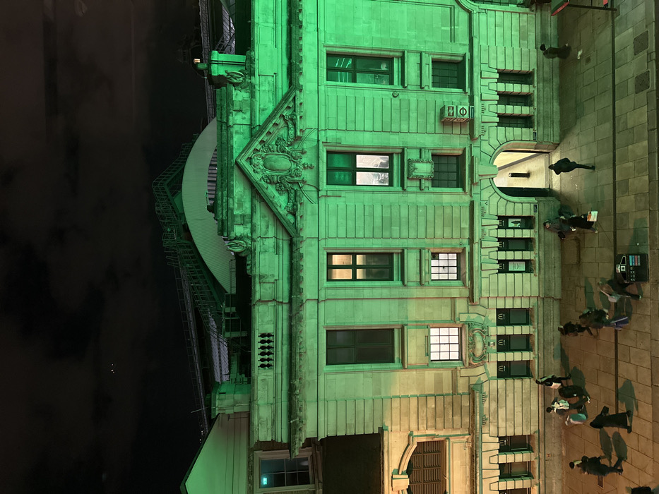 An imposing building, illuminated with vibrant green light, dominates the scene. Its facade features rows of windows, some lit, and ornate architectural details. To the left, a curved structure with an external staircase rises against a dark sky. Along the right side, a sidewalk bustles with pedestrians, their figures foreshortened from the elevated perspective. A payphone stands among the people. The overall impression is of a vibrant urban environment at night.