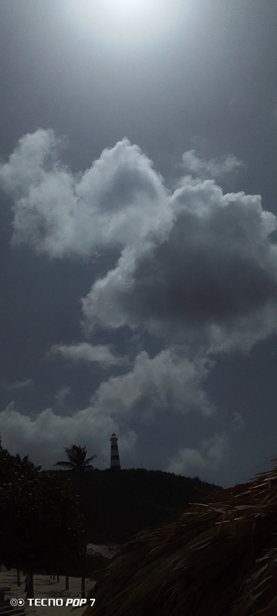 The image shows a cloudy sky above a landscape in Pedro González, Venezuela. A black-and-white striped lighthouse stands on a hill amidst trees. In the foreground, there are dark silhouettes of vegetation, possibly trees or bushes, and the edge of a thatched roof structure. The sky is overcast with thick, gray clouds. The sun is faintly visible. The scene is dark and lacks vibrant colors. "Tecno Pop 7" is watermarked at the bottom left of the image.