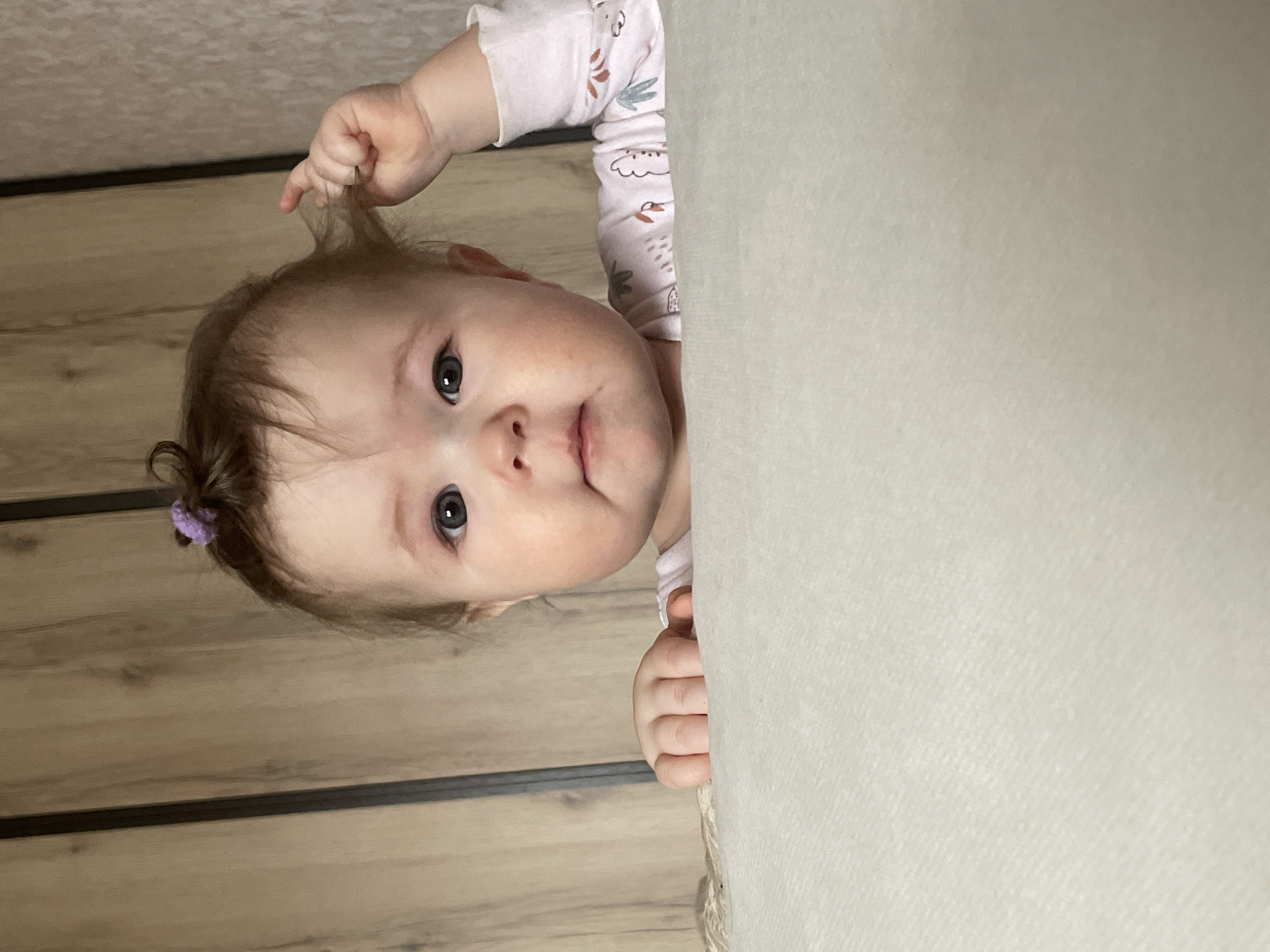 A baby peers into the frame from above a light-colored, possibly textured, surface. The baby has fair skin, light brown hair with a small purple hair tie on the left side, and is wearing a long-sleeved shirt with a pattern. The baby's right hand is holding a strand of hair, while the left hand is gripping the edge of the surface. The background consists of a wall with a wood-like pattern. The image location is presumed to be in a residential interior in Chaikovsky, Russia.