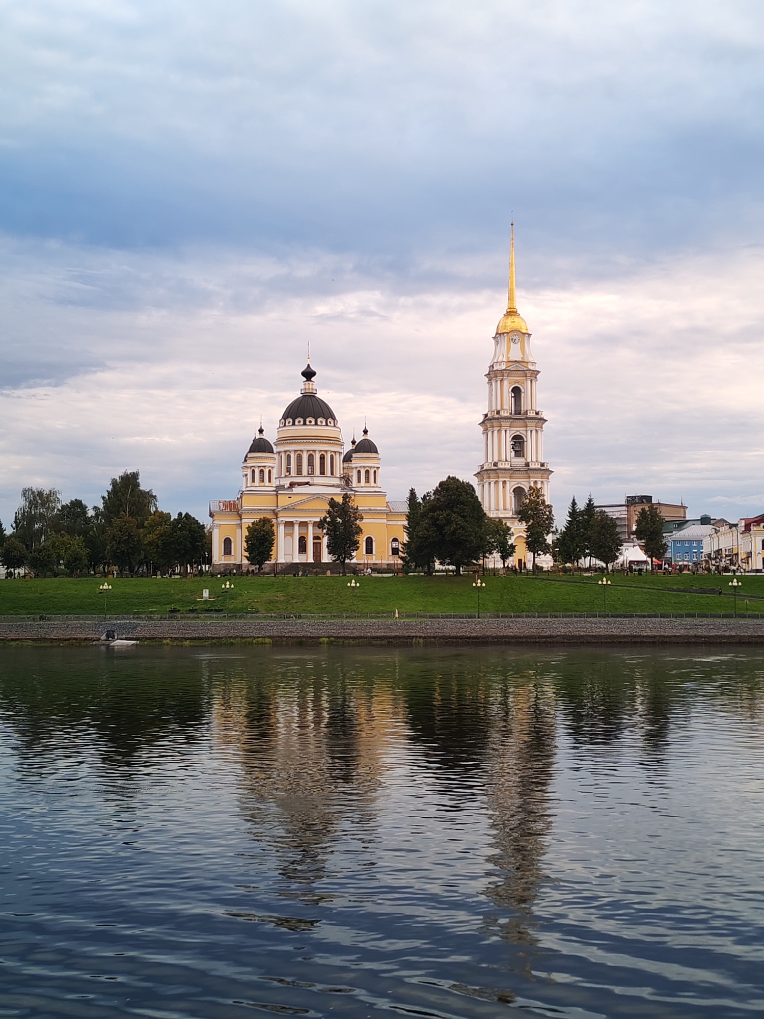 A wide shot captures a cathedral and its bell tower reflected in the calm waters of a river, with a cloudy sky overhead. The yellow cathedral has multiple domes topped with black, and the bell tower is tall and ornate, with a golden spire. Green grass covers the embankment where the cathedral sits, and trees are scattered throughout the scene. In the background, buildings line the shore. No people are visible in the image. The image appears to be a photograph taken from across the river, likely in the city of Bol'shie Mkhi, Russia, given the architectural style which is characteristic of Russian Orthodox cathedrals.