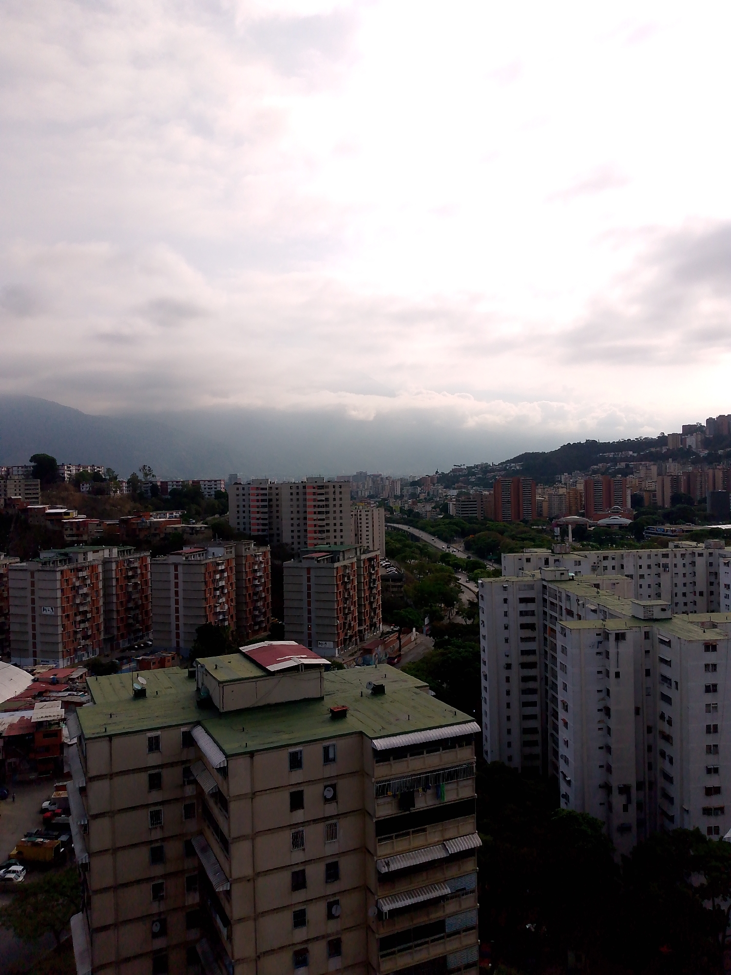 The image presents a wide, elevated view of a dense urban landscape in Caracas, Venezuela, captured on an overcast day. The sky is dominated by thick, grey clouds that diffuse the sunlight, lending a soft, muted quality to the scene.
The primary subjects are numerous high-rise residential buildings, predominantly apartment blocks, that stretch across a hilly terrain. These buildings vary in appearance; many feature balconies, some with retractable awnings, and visible window patterns. A common architectural theme includes structures with reddish-brown or exposed brick-like facades, alongside others painted in lighter neutral tones. Several closer buildings exhibit distinctive green-colored flat roofs.
In the background, mountains rise majestically, their upper slopes and peaks partially obscured by the low-hanging clouds and atmospheric haze, characteristic of a cloudy day. Lush green vegetation and trees are interspersed throughout the cityscape, lining roads and occupying spaces between buildings, providing natural contrast to the concrete structures. A multi-lane road or highway is visible winding through the mid-ground, indicating a busy urban infrastructure.
No specific human activity or event is distinctly captured, though the sheer number of residential units implies a bustling city life. A few parked cars can be seen near the bases of some buildings. The time of day appears to be daytime, likely late morning or afternoon, given the overall illumination despite the heavy cloud cover. The weather is clearly cloudy and overcast. No legible text is visible within the image.