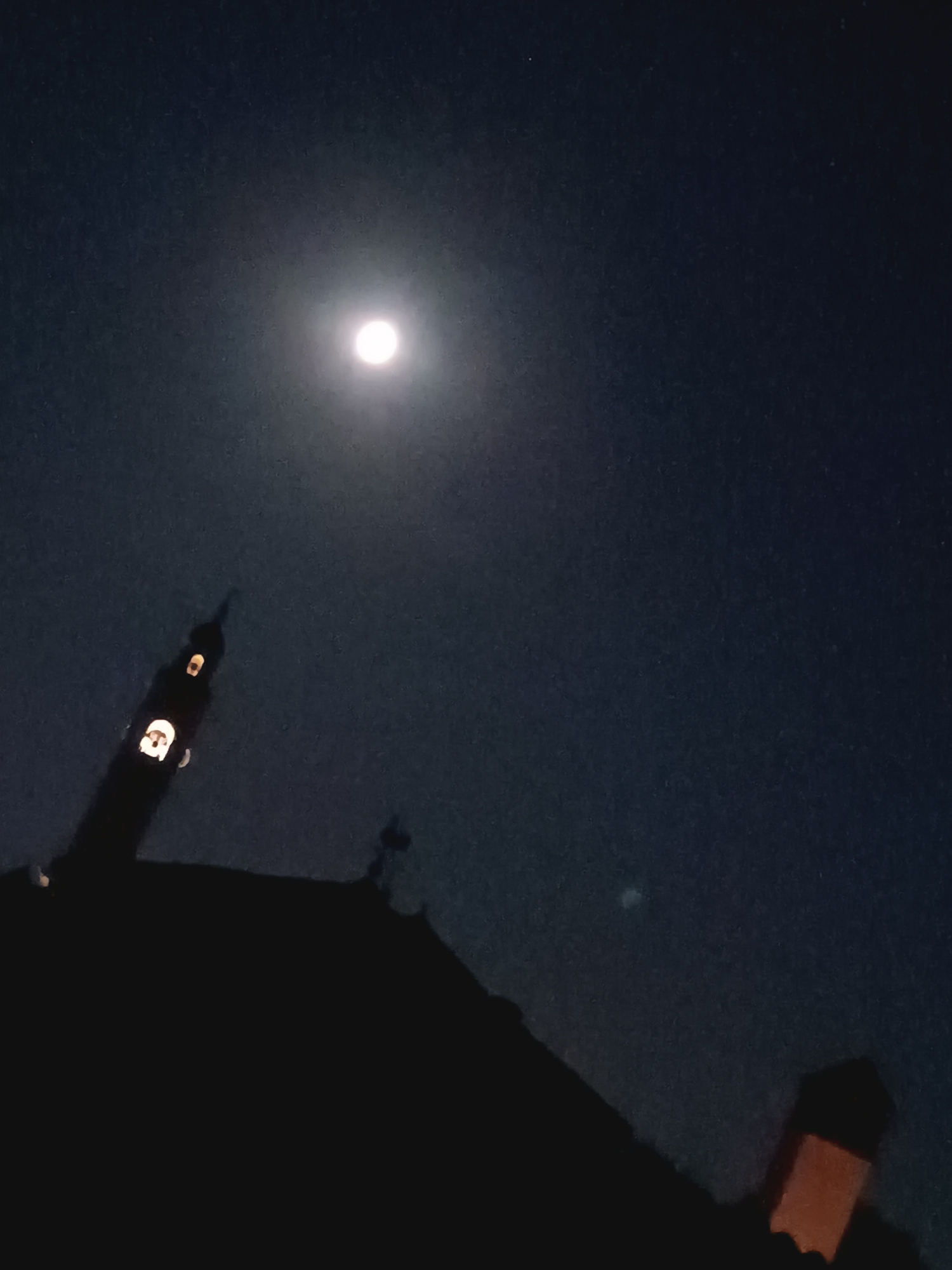 The image shows a nighttime view of a town, with a bright moon in the dark sky. Silhouetted against the sky is a prominent tower with illuminated windows. A portion of a building's roof is visible in the foreground, also in silhouette. The context of Viadana, Italy is provided, suggesting the location of this scene.