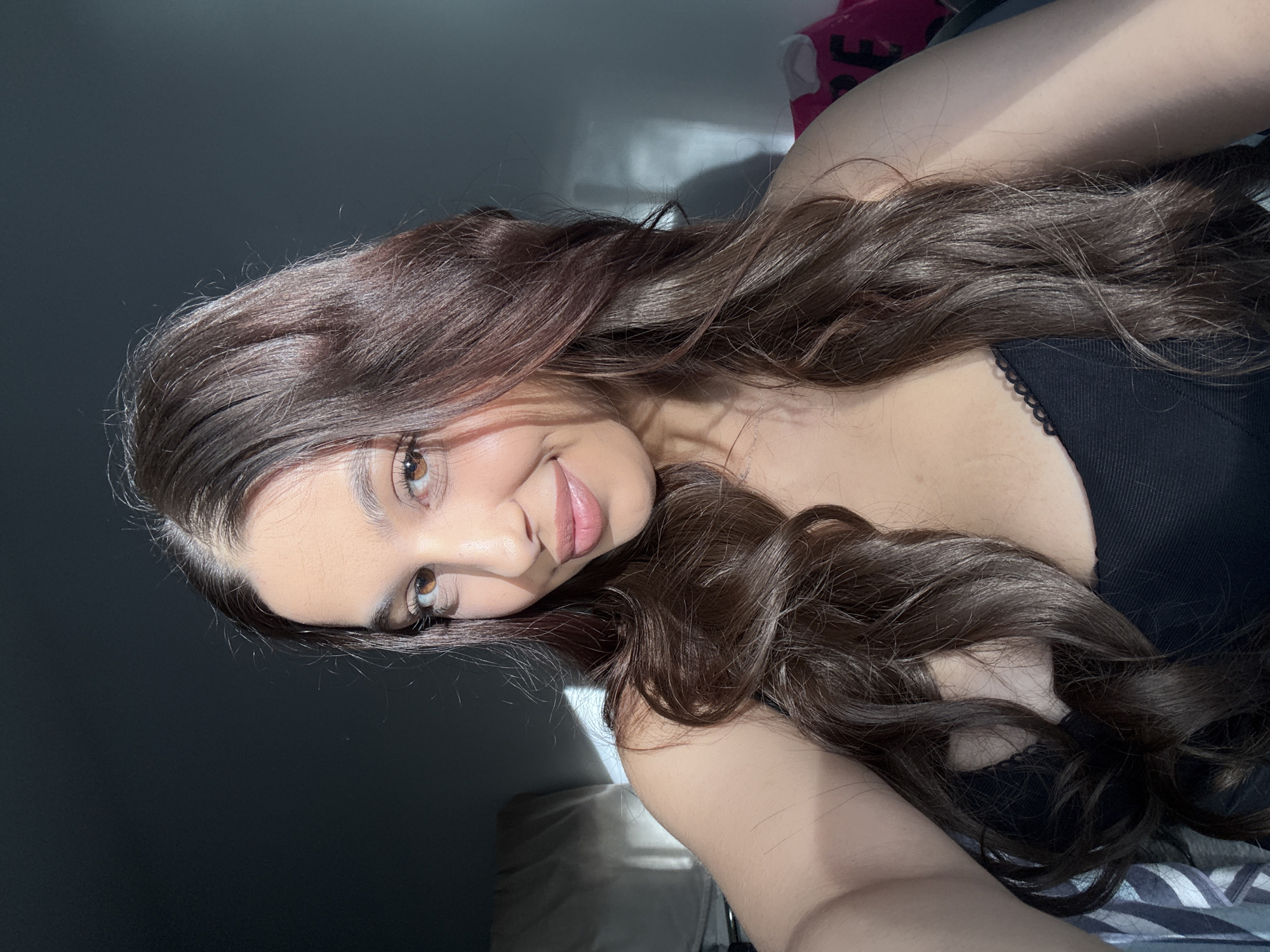 A young woman is lying on her side in an interior setting. She has long, wavy brown hair and fair skin, wearing a black top. The background is blurred, with a grey wall visible behind her. The location is not identifiable from this image as being in Mönchengladbach, Germany.