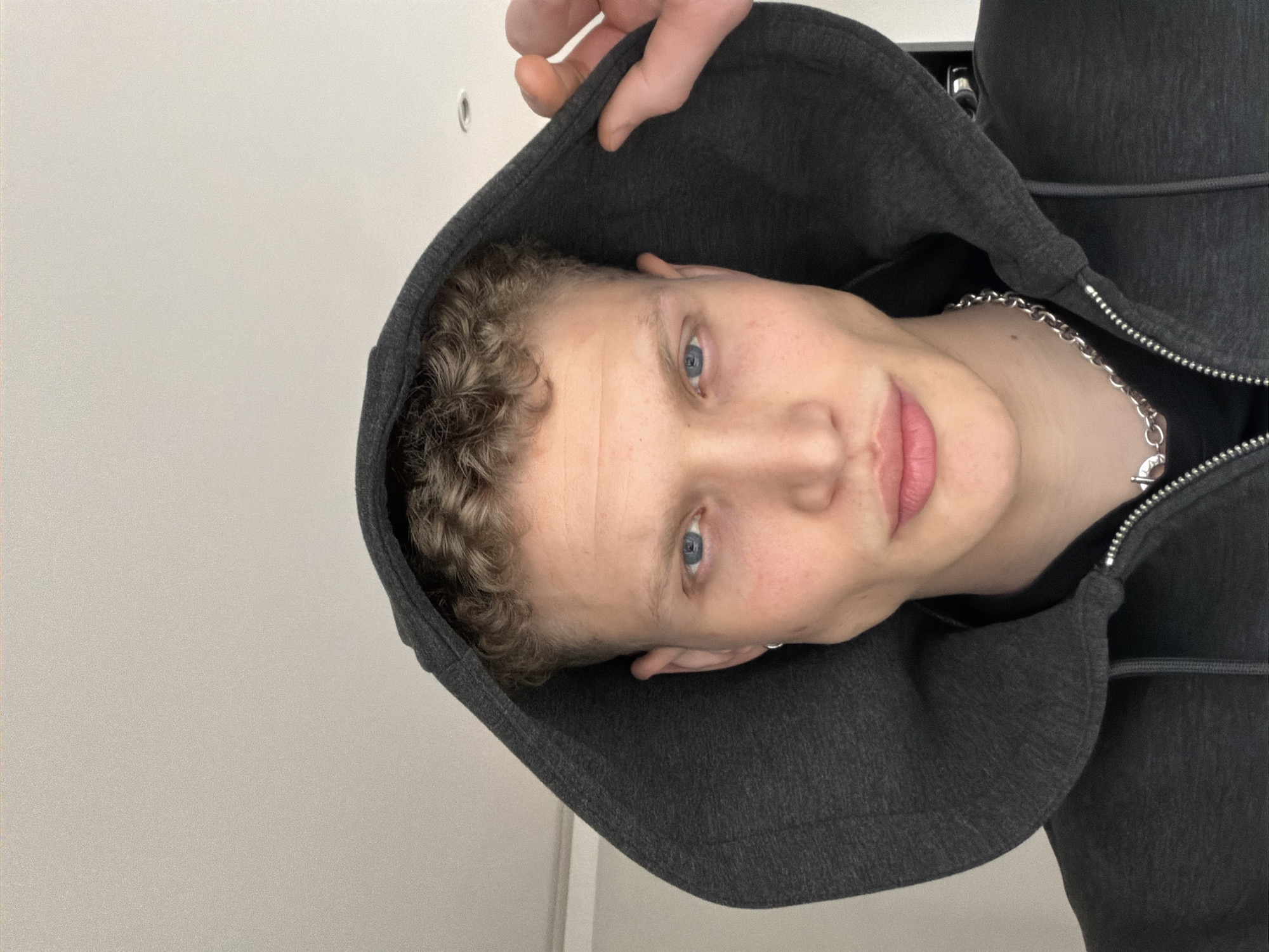 A person with light skin and short, curly blond hair is wearing a dark gray hooded sweatshirt. The person is facing the camera, with a slight smile. The hood is partially up, obscuring the top of the head. A silver chain necklace is visible around the neck. The background is a plain white wall. The location is not discernible from the image.