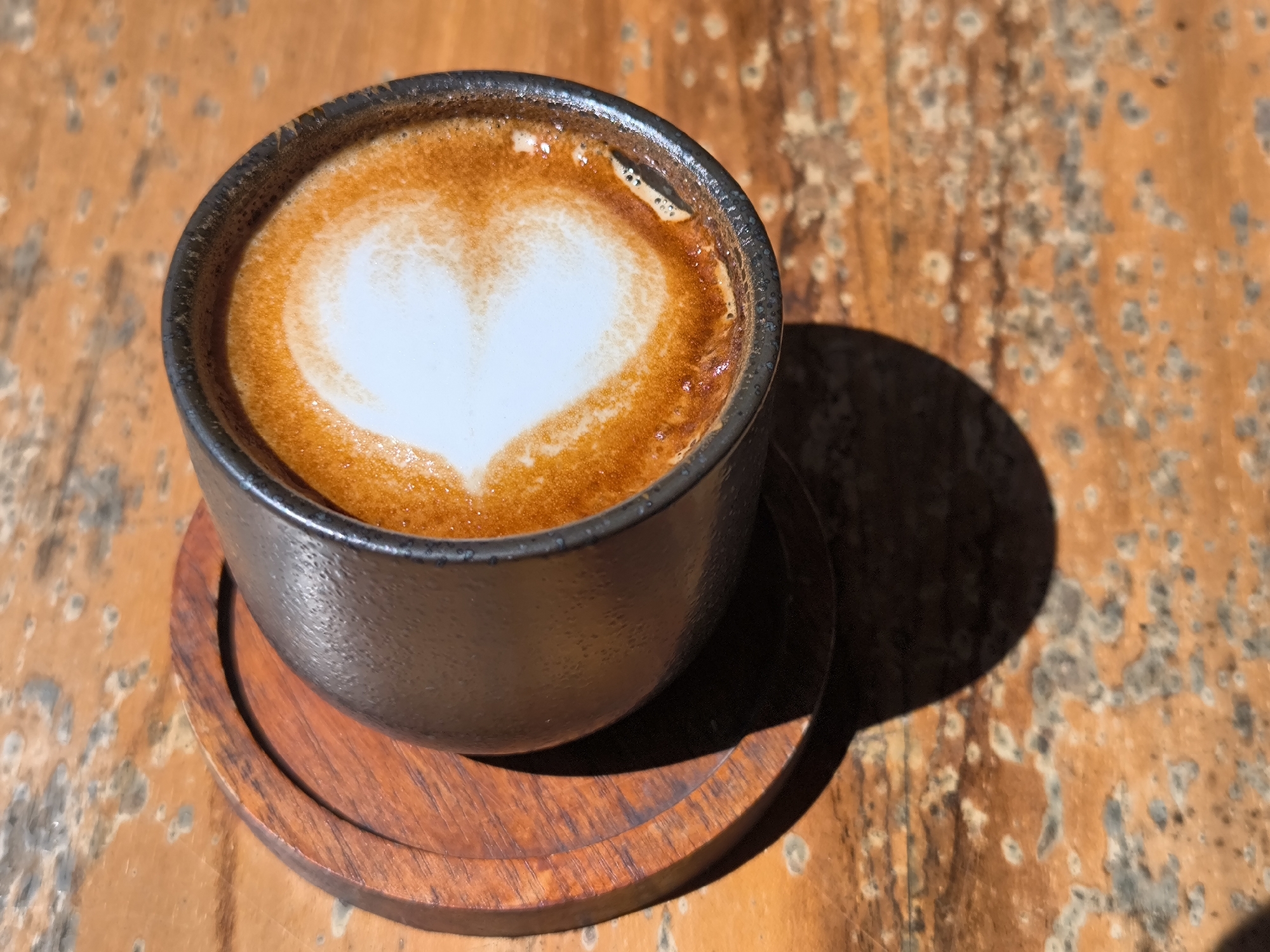 A close-up shot shows a black, round ceramic cup filled with a cappuccino, featuring latte art in the shape of a heart. The cup rests on a dark-stained wooden coaster on top of a weathered wooden table. The table exhibits an orange-brown color with a mottled pattern, showing signs of wear and age. A shadow cast by the cup and coaster extends to the right of the objects, indicating a light source from the left. The photo was taken in the city of Sembalun Bumbung, Indonesia.