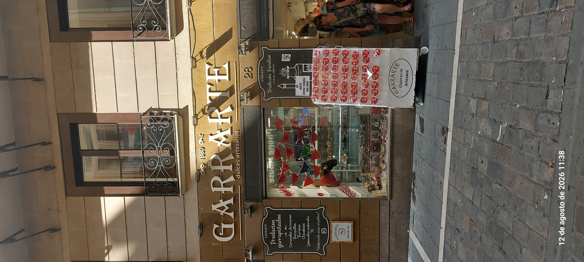 The image shows the exterior of a candy shop named "GARRARTE" in Pamplona, Spain. The shop has a large display window filled with sweets and a prominent sign with the shop's name in white lettering. To the right of the entrance, a cart displays rows of red lollipops. A woman wearing sunglasses and a patterned dress is walking towards the shop on the right side of the frame. The street in front of the shop is paved with cobblestones. A timestamp at the bottom of the image reads "12 de agosto de 2026 11:38".