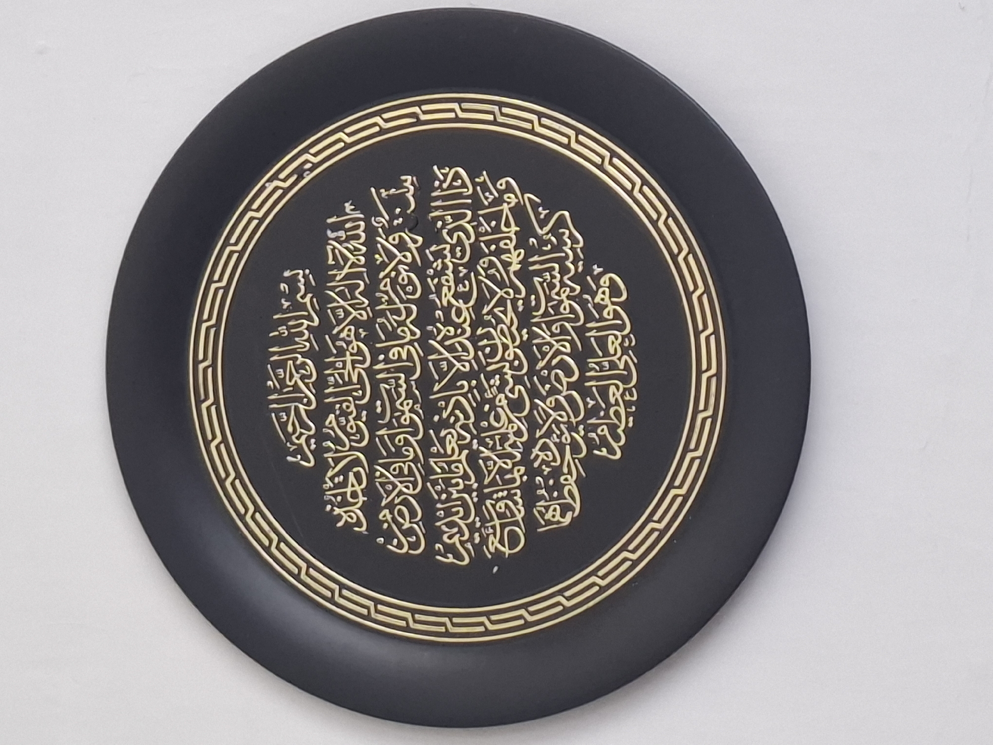 This is a close-up, eye-level shot of a round, black decorative plate with gold Arabic calligraphy. The plate is centered in the frame and appears to be hanging against a plain white background, suggesting it is either indoors or against a white wall. The calligraphy is intricate and prominent, filling the central circular design of the plate. The plate itself has a raised, patterned border in gold that frames the calligraphy. The lighting is even, highlighting the texture and details of the plate and its inscription. There are no people or any other objects visible in the image, and no contextual clues suggest a specific location in Almaty, Kazakhstan.