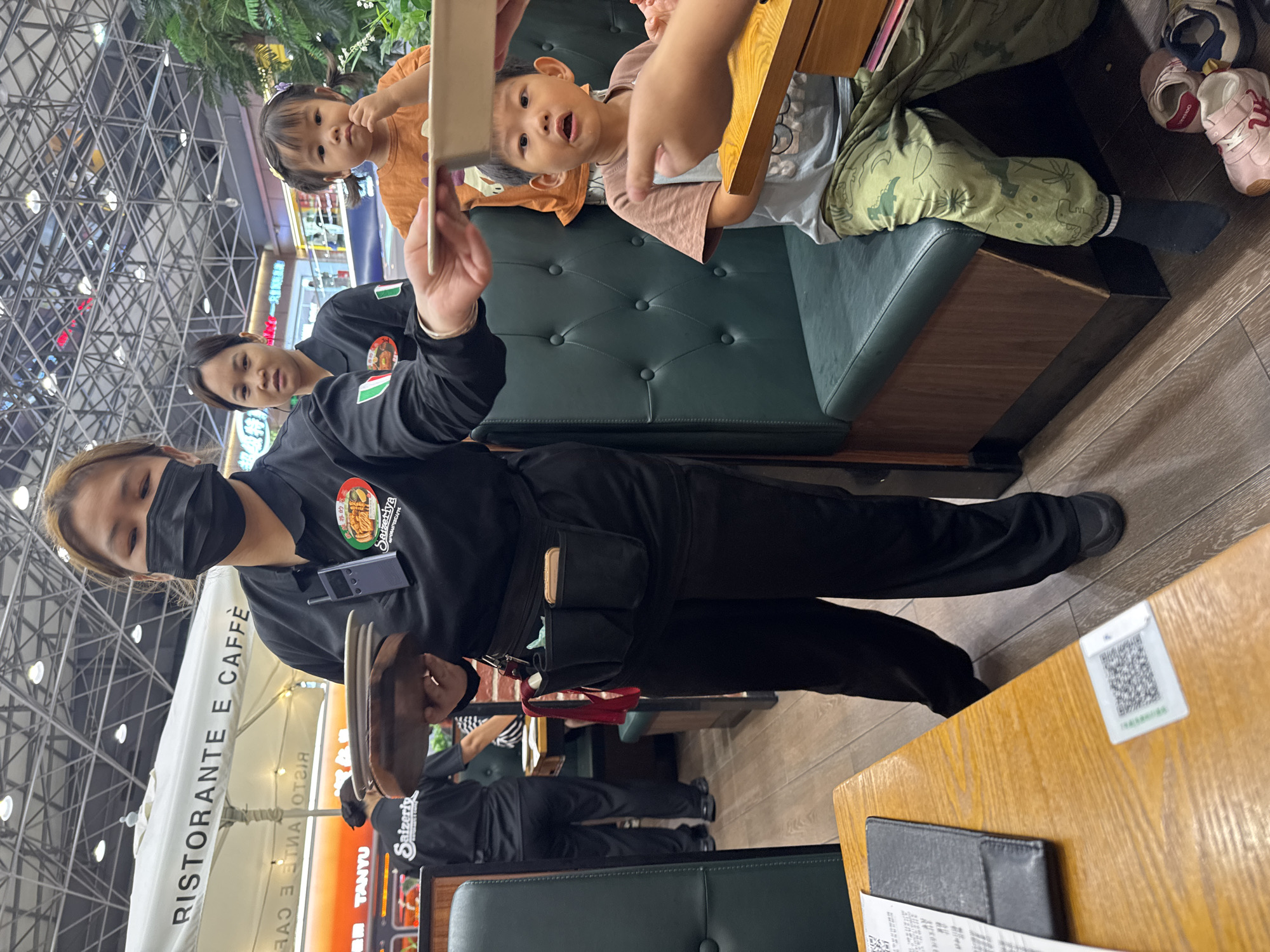 A waiter wearing a black uniform and a black face mask holds a tray while serving children seated on a green leather booth at Ristorante E Caffè in Haizhu District, China. The children are looking at the waiter and the tray. The surrounding environment includes a restaurant interior with tables, chairs, and an outdoor area visible through the window. The booth seating includes an occupant wearing patterned pants and shoes near the side of the seat.