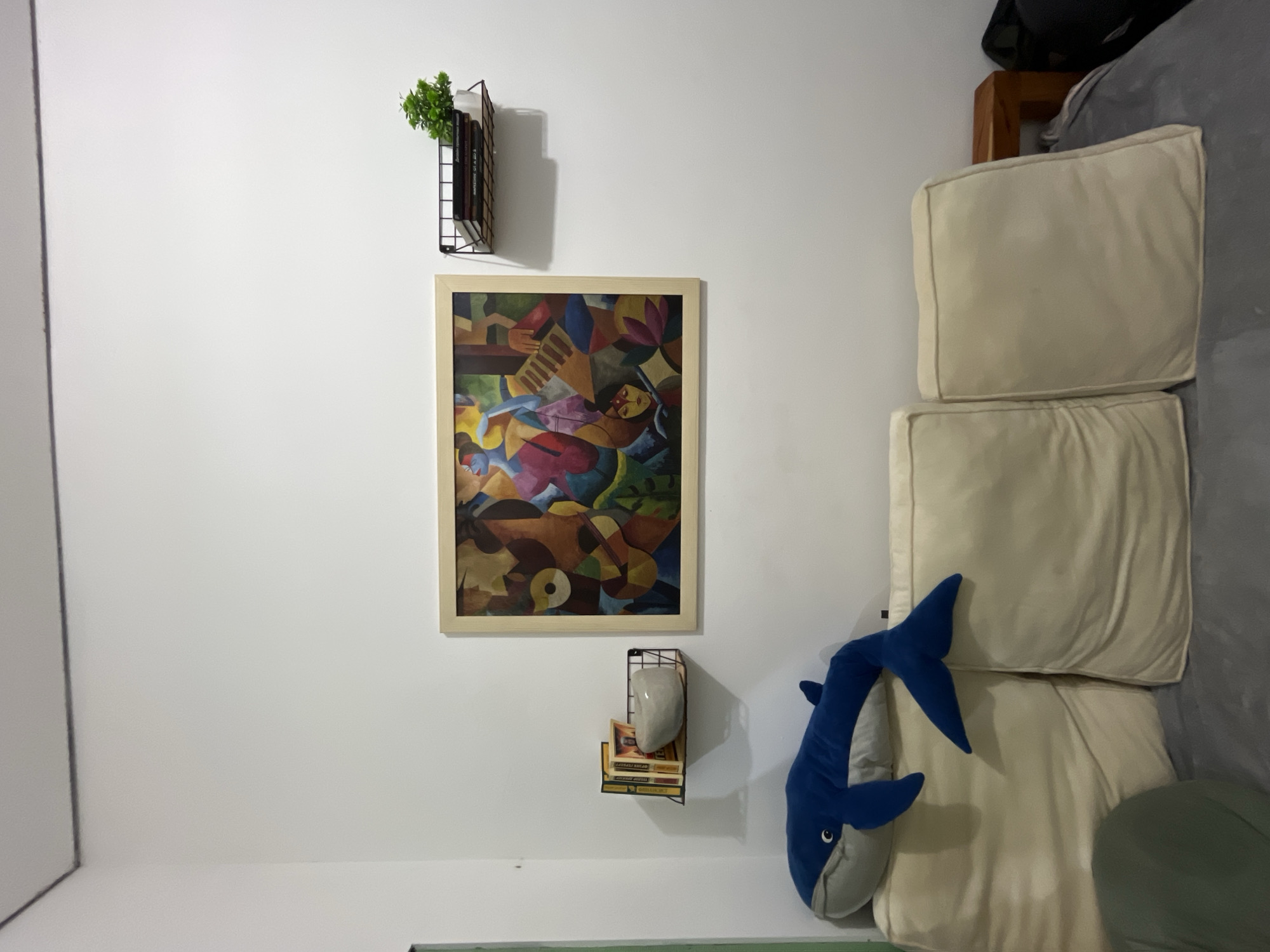 The image displays an indoor scene, likely a living room or bedroom, with a white wall adorned with artwork and shelving, and a sofa on the right.
Dominating the center of the white wall is a framed abstract painting. The artwork features a vibrant composition of geometric shapes and stylized figures in various shades of brown, orange, blue, green, pink, and yellow, with one figure seemingly playing an accordion. Above the painting, a black wire wall-mounted shelf holds a small artificial green plant and a few books, whose titles are not discernable. Below the painting, another black wire shelf supports several books and a light gray, irregularly shaped decorative object. On one of the books, text is partially visible, reading "LEGACY AHEAD" and "THE GOLDEN".
To the right, a sofa or bed with light cream-colored cushions is visible, covered with a grey fabric. Resting on these cushions is a large, blue shark plush toy, positioned with its head pointing towards the left of the frame. A wooden structure, possibly a bed frame or headboard, is partially seen at the top right corner behind the cushions. A hint of a green cushion is also visible at the bottom right. The scene is brightly lit by ambient indoor light, depicting a neat and decorated interior, consistent with a home setting in Kuta Selatan, Indonesia.