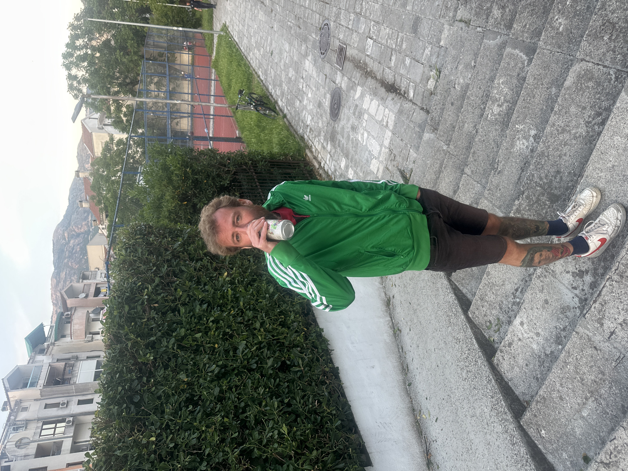 A man stands on concrete steps on a sidewalk in Budva, Montenegro. He is drinking from a can. He wears a green Adidas track jacket, brown shorts, and white sneakers with red accents. He has tattoos on his lower left leg. Behind him are buildings and a hill/mountain. To his left is a large bush and to the right is a paved path leading away from the steps. In the background is a fenced sports court.