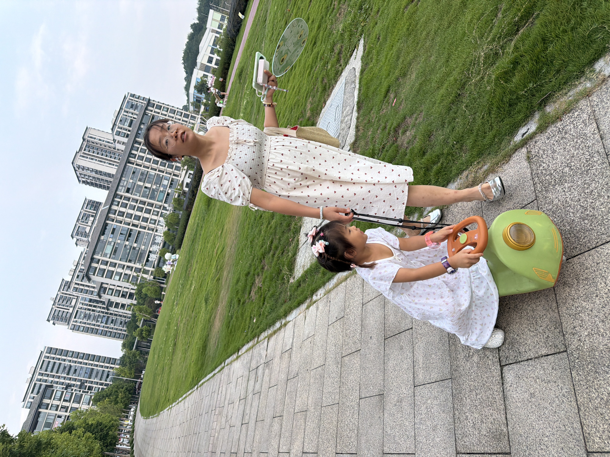 A woman and a young girl are in a park area in Jiangjin, China. The woman, wearing glasses and a white dress with red heart patterns, stands on a paved walkway, holding a fan and a phone. The young girl, also in a white dress, sits on a green toy car with an orange steering wheel, gripping its handlebars. The park features a large grassy area and a row of modern apartment buildings in the background. The sky is overcast.