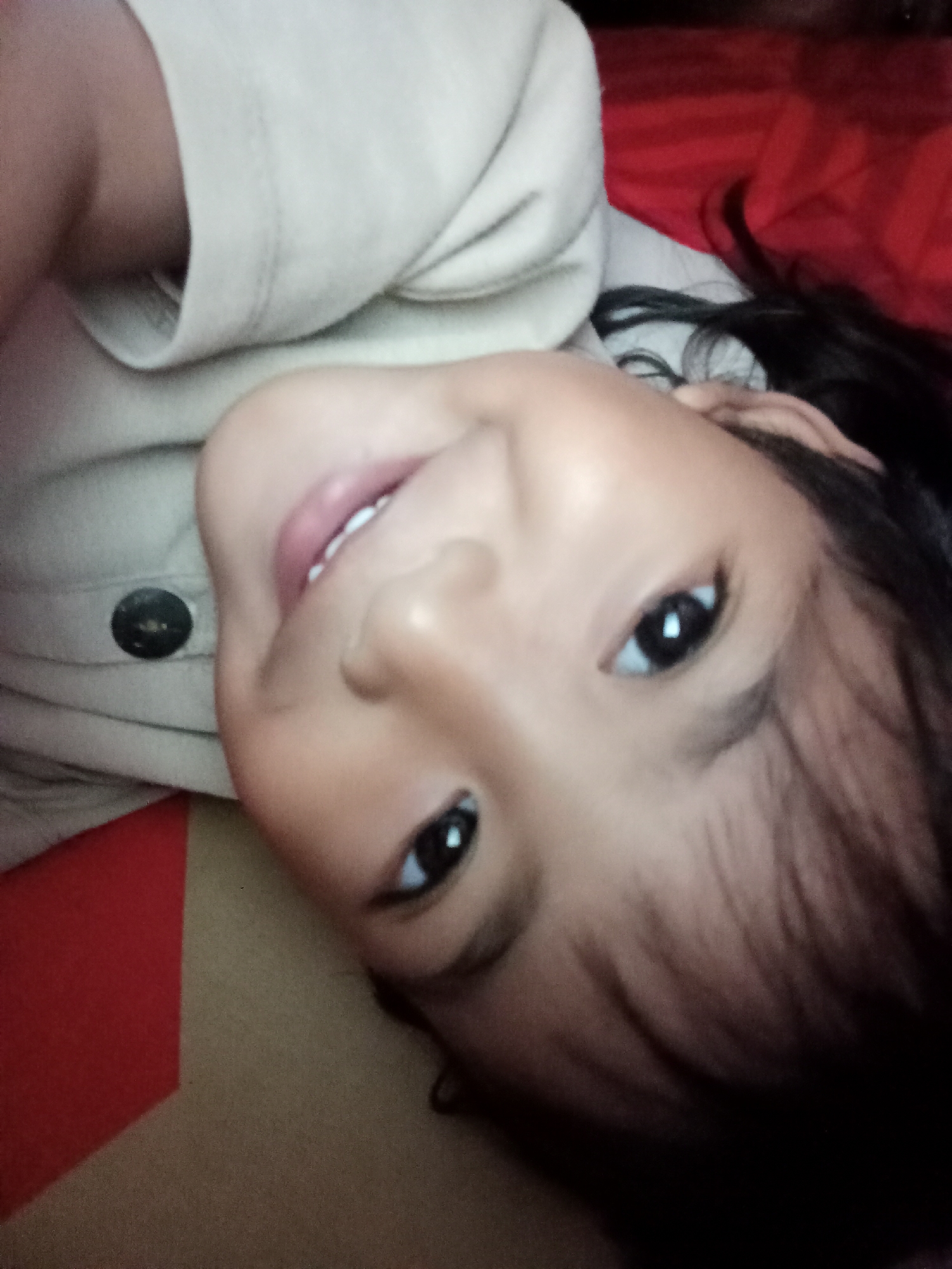 This is a close-up, sideways, low-angle, slightly blurry photograph of a young child lying down. The child has fair skin, dark hair, and is wearing a light-colored shirt with a dark button. The child is looking directly at the camera with a slight smile, showing teeth. The background is indistinct, but appears to include a red surface and a brown surface. The image is oriented such that the child's head is to the right and their feet are to the left. There is no discernible location in Bloro, Indonesia.