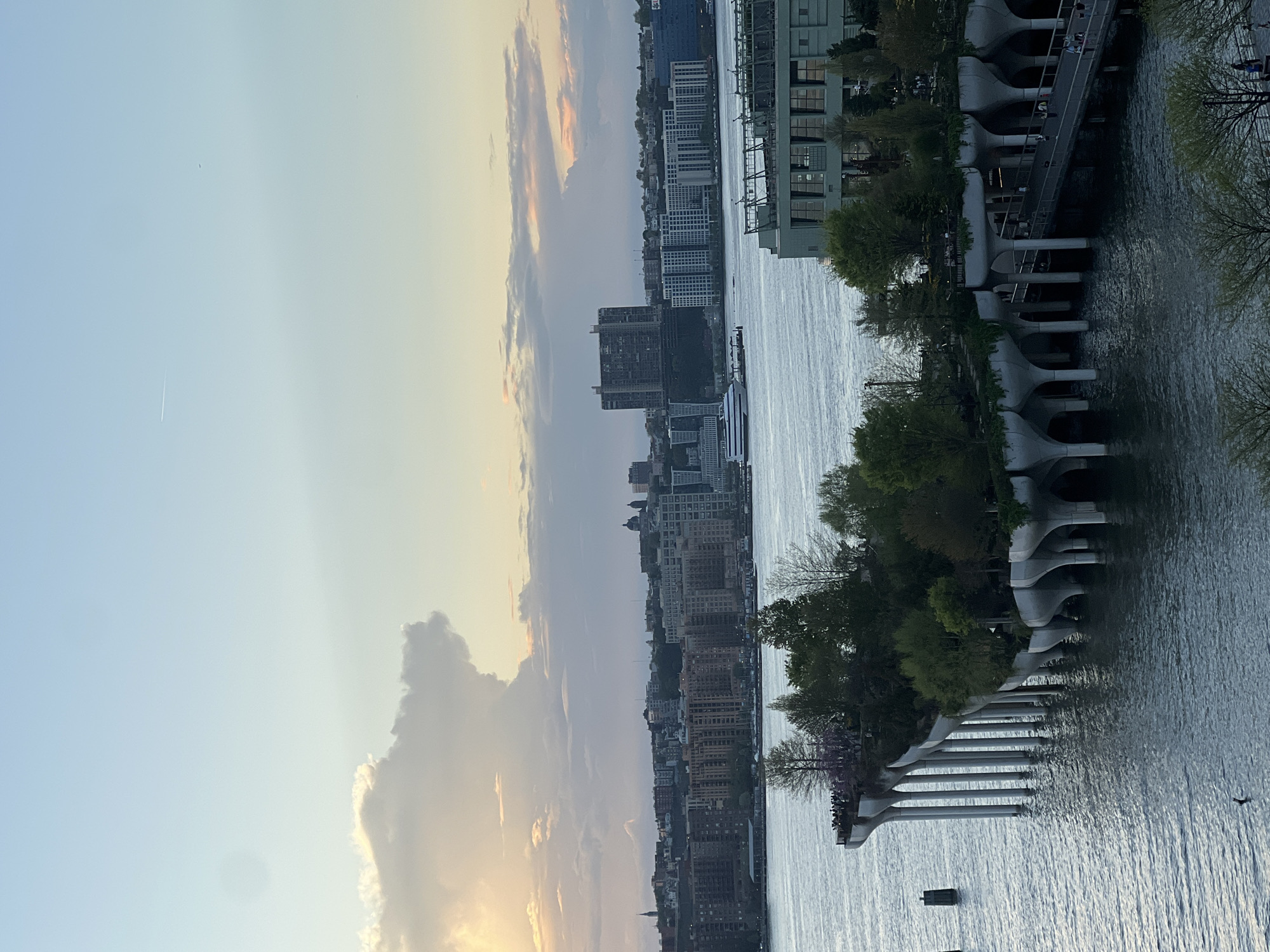 This photograph depicts a cityscape along a body of water, likely in New York City, United States. The sky is characterized by clouds and the light from the sun. Buildings of varying heights are visible along the waterfront. There is a structure with a unique architectural design, possibly a pier or public space extending into the water. The water surface shows light reflecting off it.