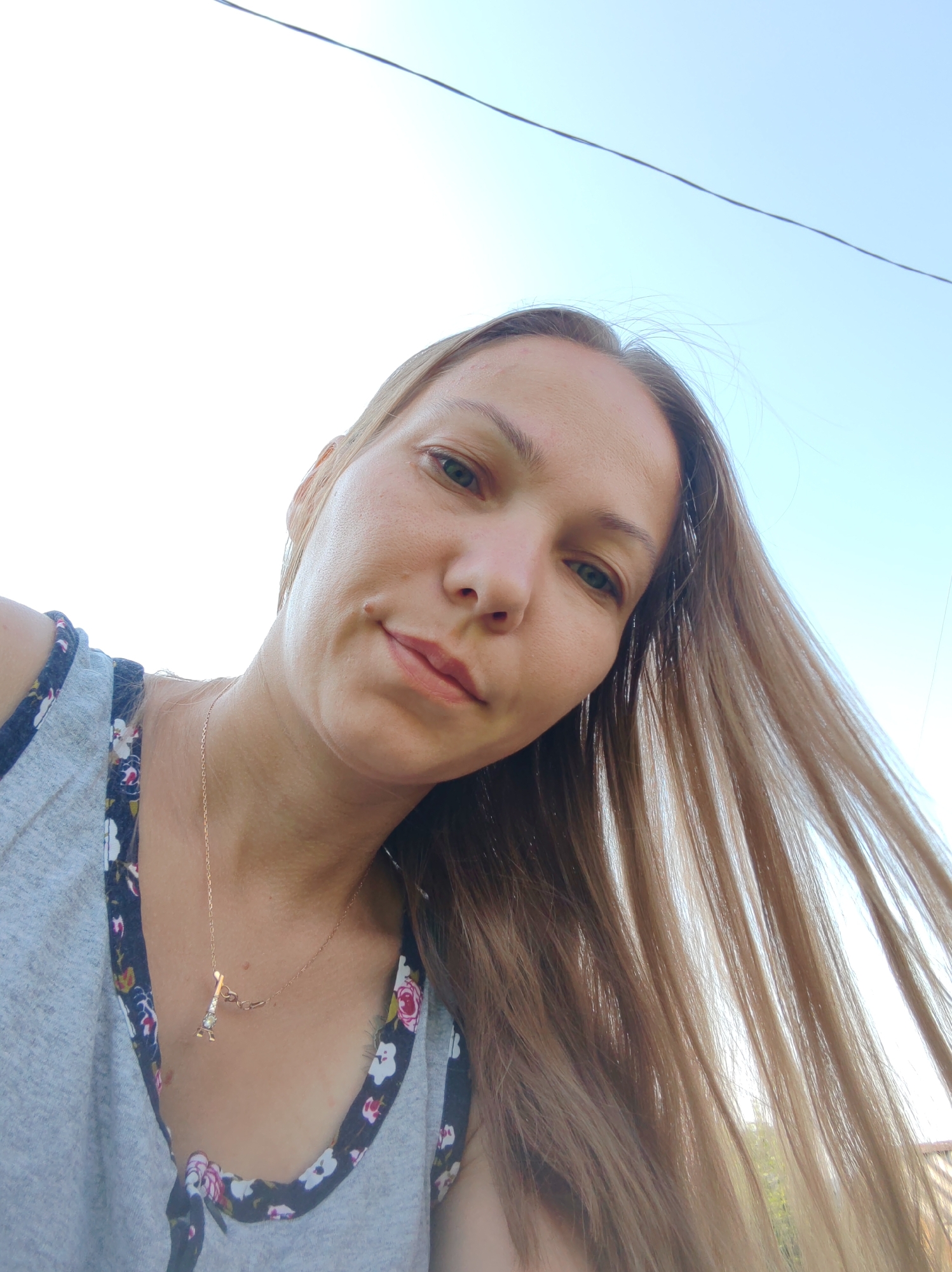 The image shows a close-up, low-angle, outdoor selfie of a young woman with long, light brown hair. She is wearing a grey tank top with a floral trim. A delicate gold necklace with a small Eiffel Tower pendant is visible around her neck. The background consists of a bright, clear sky and a thin black wire stretching across the top of the frame. There are hints of greenery and a building in the far distance. The lighting suggests it is daytime. The setting is likely a residential area in Чухлэм, Russia, given the context.