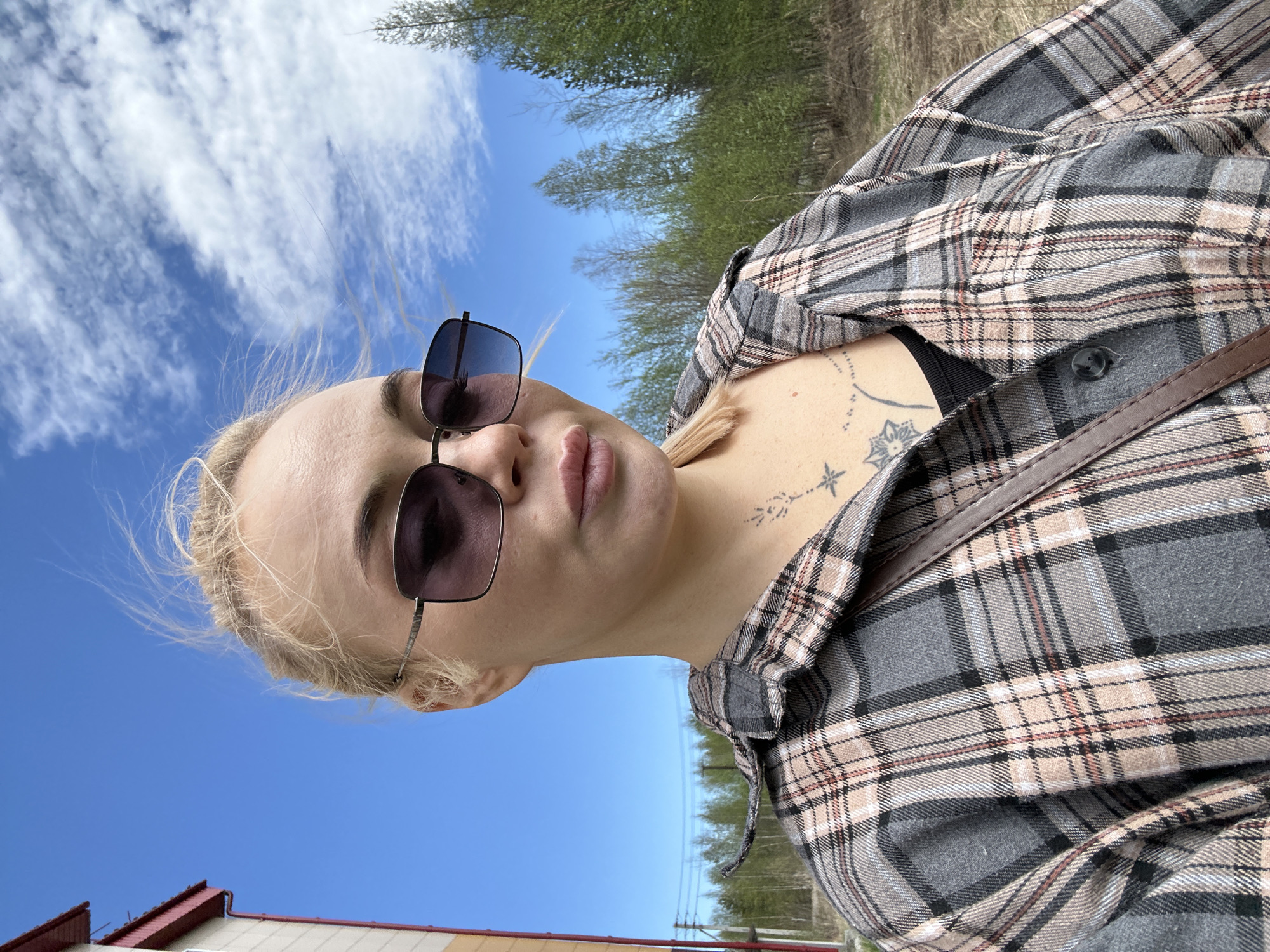 A woman with blonde hair and sunglasses is the main subject of the outdoor selfie. She is wearing a grey plaid shirt with a brown strap across her torso. A tattoo is visible on her chest. The background features a blue sky with scattered clouds, trees, and the roof of a building. This image was likely taken in the city of Высокий, Russia.