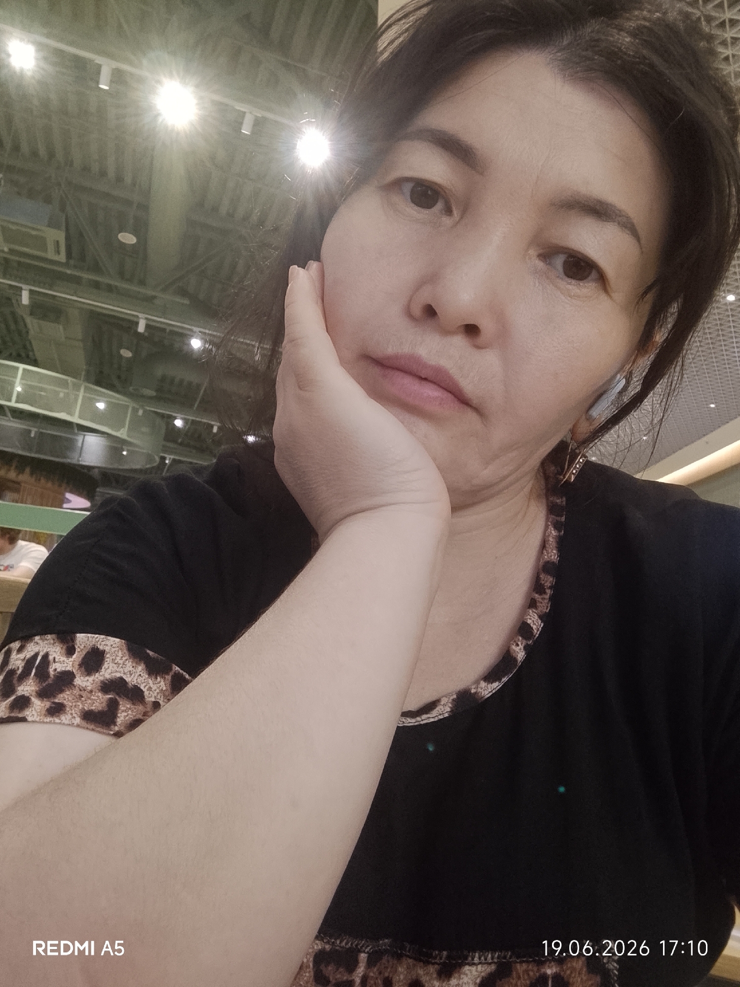 A woman is captured in a close-up selfie, resting her chin on her hand. She has dark hair, dark eyes, and fair skin. She is wearing a black t-shirt with a leopard print trim around the neckline. The background appears to be indoors, with a ceiling featuring exposed metal beams and fluorescent lighting. There are some blurry elements in the background suggesting a public space, possibly a mall or restaurant, with other people and tables visible. The overall lighting is bright, with some lights creating a starburst effect. The timestamp at the bottom right indicates the photo was taken on June 19, 2026, at 17:10. The text "REDMI A5" is also visible in the bottom left corner, likely indicating the phone model used to take the picture. The woman's expression is neutral, with a hint of thoughtfulness as she gazes towards the camera.