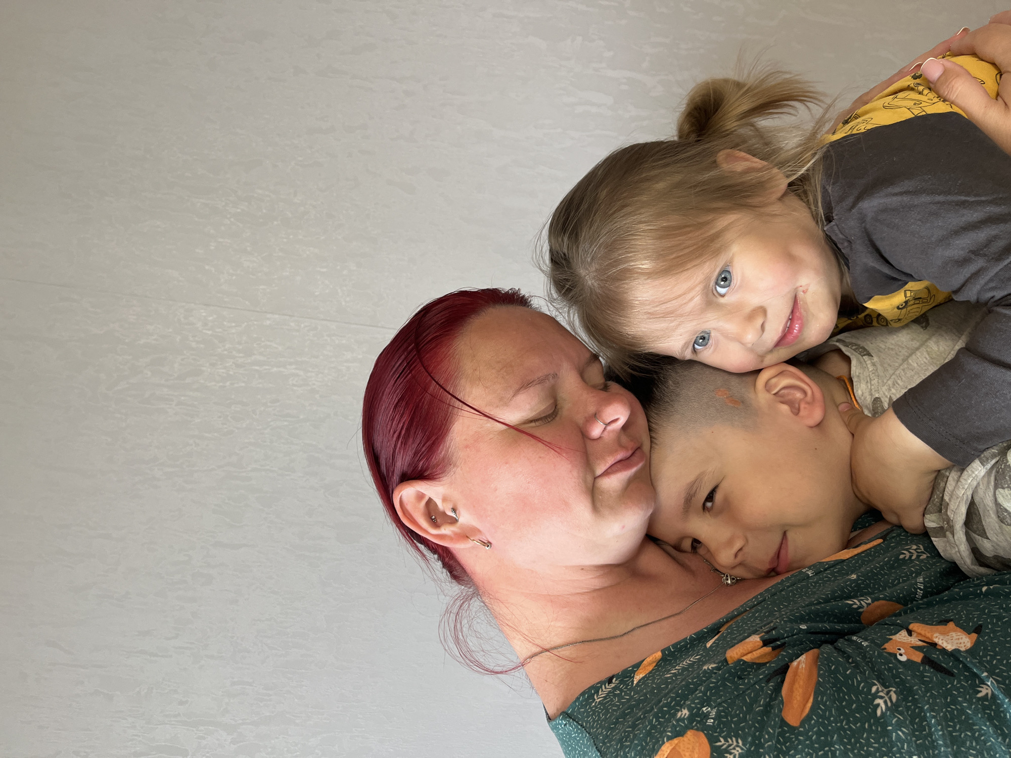 A woman with red hair is embracing two children. One child, a young boy with short hair, is lying on her chest, his head turned towards her. The other child, a young girl with blonde hair in a ponytail, is on top of the boy, her head close to the woman's. The woman's eyes are closed. The scene appears to be indoors, with a white wall in the background. There is no clear evidence of it being located in Chaikovsky, Russia.