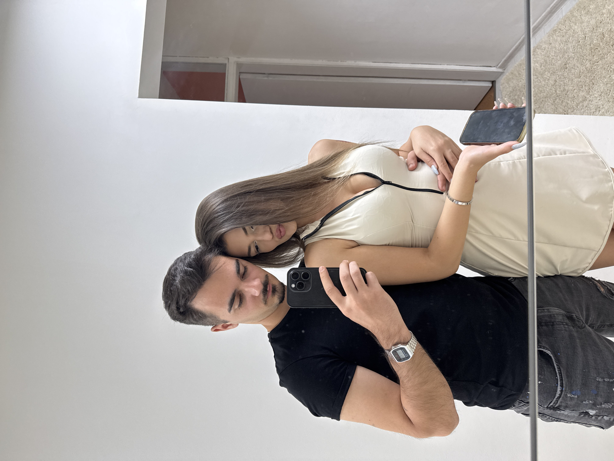 A young man and woman are posing for a mirror selfie in a well-lit hallway. The man, on the left, wears a black t-shirt and distressed grey jeans and has a watch on his left wrist. He holds a black smartphone in his right hand, angled to capture their reflection. The woman, on the right, leans into him, wearing a sleeveless cream-colored dress with a black zipper down the front. She holds another black smartphone in her left hand. Their reflections are visible in a large mirror that spans the lower portion of the frame. The background shows a white wall and a doorway leading to another room. The environment suggests an indoor residential or commercial space, potentially in Sofia, Bulgaria, given the context.