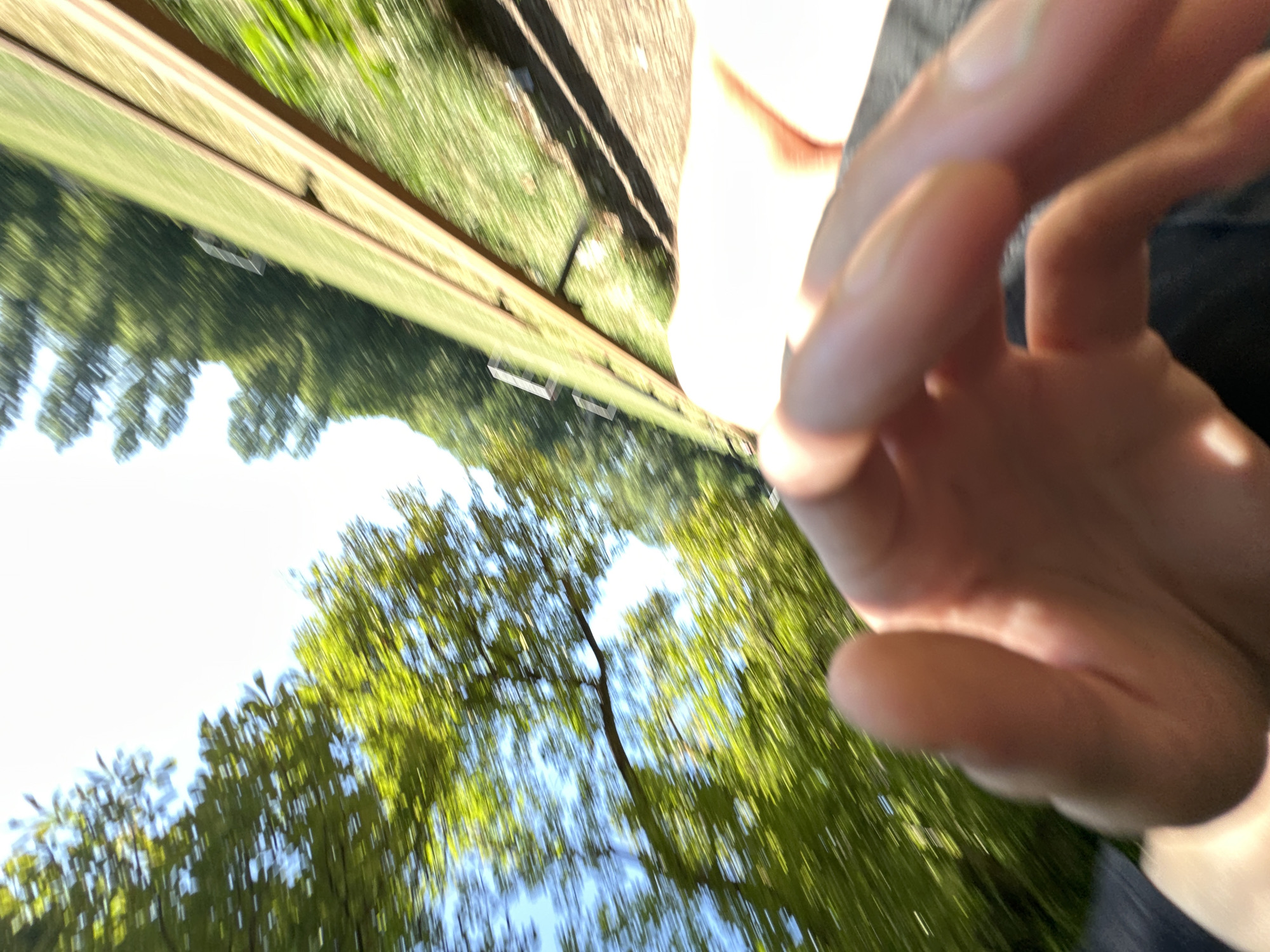 The image shows a blurred, first-person perspective of a hand reaching out towards a grassy area next to a railway track. Trees with green foliage are visible in the background, along with a bright sky. The motion blur suggests movement, possibly from a vehicle or the person holding the camera. There is no specific indication of the location being the city of Makiv, Ukraine.