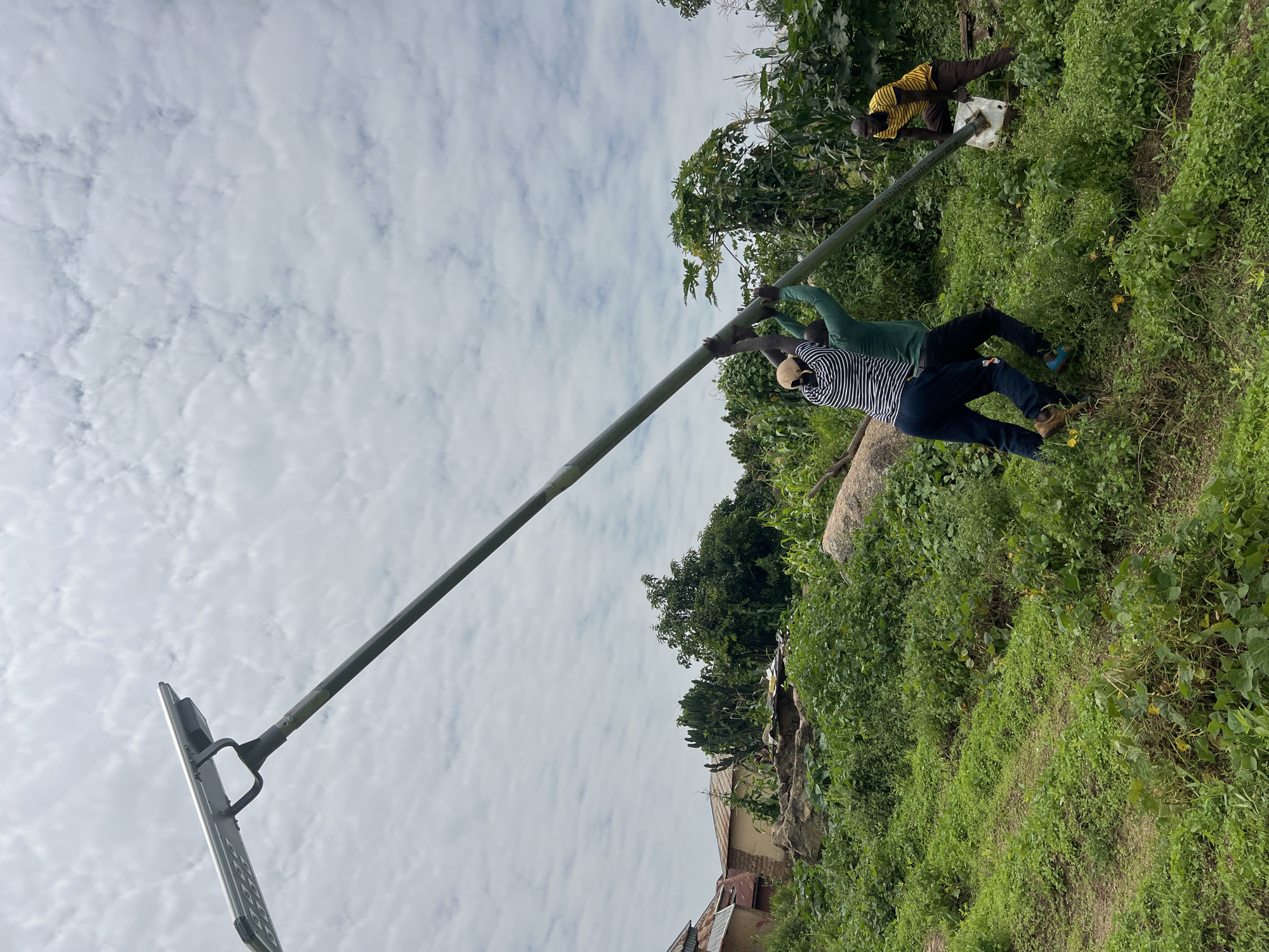 In Dong, Nigeria, four men are observed working together to install a solar-powered street light. The scene unfolds on a grassy slope dotted with rocks and vegetation. The primary action involves three men lifting and maneuvering a long, metallic pole with a solar panel attached at the top. Two men are at the lower end of the pole, pushing it upwards and to the left, while a third man is positioned higher up the pole, assisting with the lift. A fourth man stands to the right, holding a white bucket and observing the operation. The background features a cluster of rural buildings with tin roofs and lush green foliage, including corn stalks, suggesting a residential or agricultural area. The sky above is overcast with fluffy white clouds.