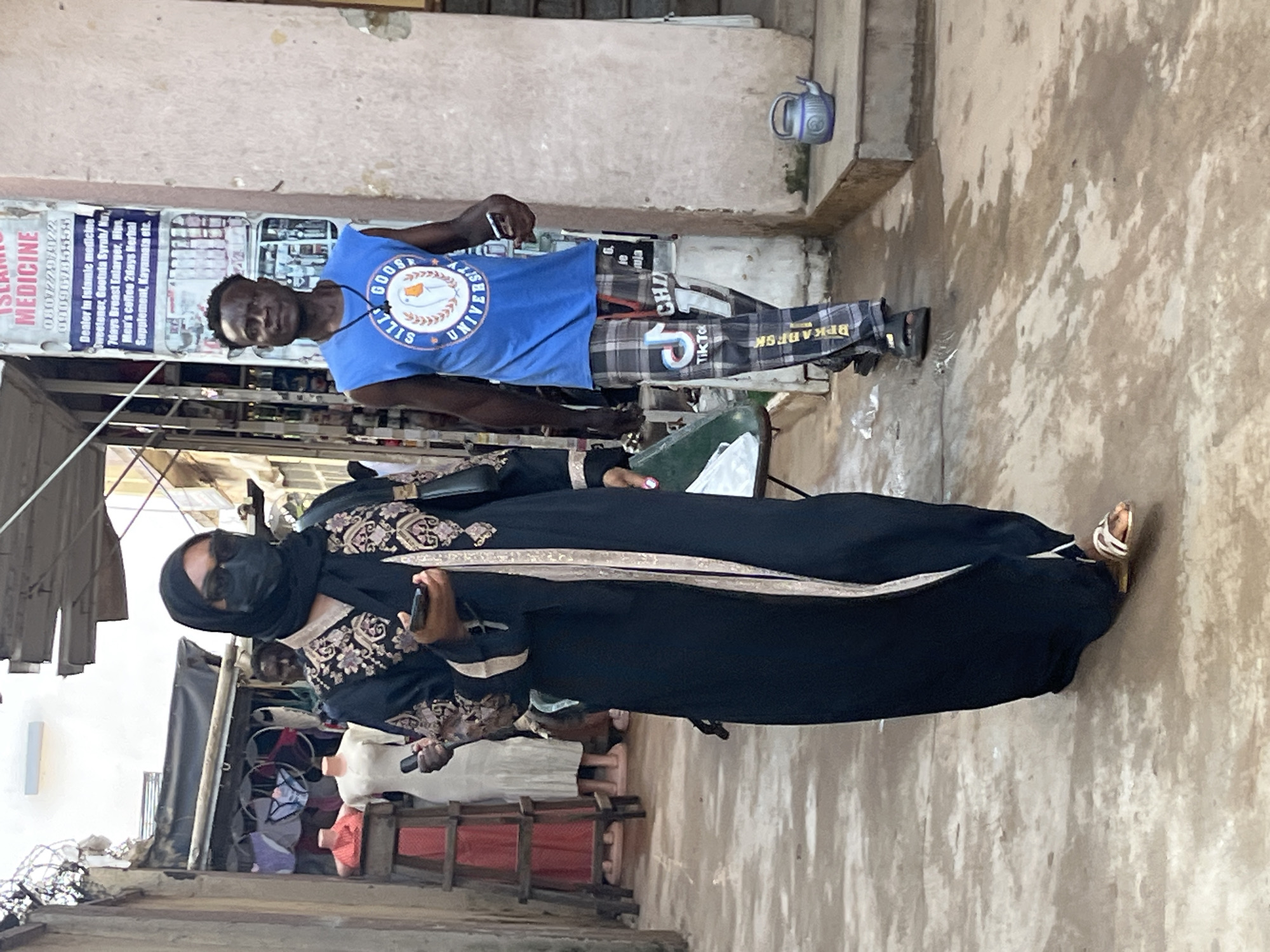 In an outdoor market alley in Abuja, Nigeria, a Black woman wearing a black hijab, sunglasses, and a black abaya with gold trim walks forward, holding a smartphone in her right hand. She is wearing sandals. To her right, a Black man wearing a blue sleeveless shirt with a "Silly Goose University" logo and checkered pants walks in the same direction, holding a smartphone in his right hand. He is wearing sandals. Behind them, on the left, mannequins display undergarments and clothing. A sign on a building behind the man advertises "Islamic Medicine" and lists various remedies. The ground is paved and appears wet in places. A blue teapot sits on a step to the right.