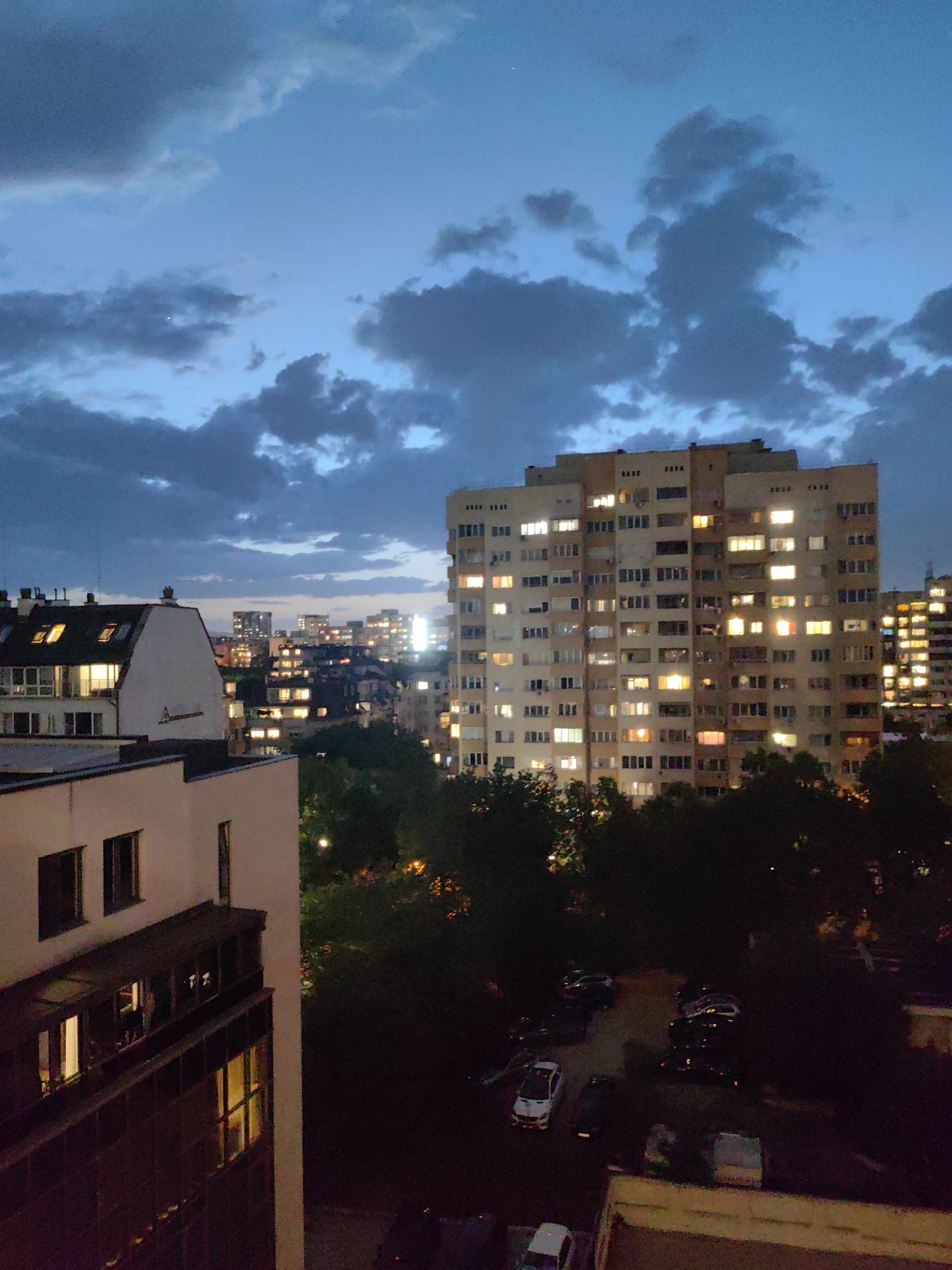 The image depicts a cityscape at dusk, likely in Sofia, Bulgaria, based on the architectural style and urban layout. A tall, multi-story apartment building with numerous illuminated windows dominates the midground. In the foreground is a shorter building with dark windows and an overhanging balcony. Green trees fill the space between the buildings, obscuring the ground level where several parked cars are visible. The sky is a deep blue with scattered, dark clouds. In the distance, more buildings with lighted windows are visible, indicating a densely populated urban area. There are no people visible in the scene.