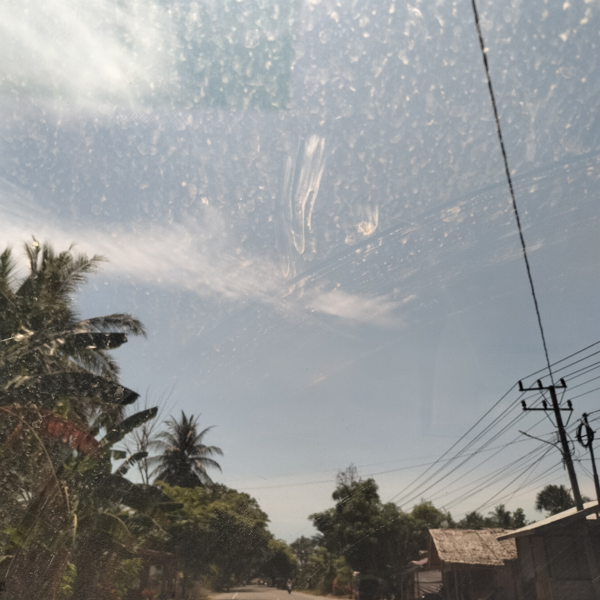 The image shows a view from inside a vehicle, likely a car, looking out at a rural landscape in Aluekeujruen, Indonesia. The windshield is dirty, with water spots and streaks obscuring the view.
Outside, the scene depicts a road lined with lush tropical vegetation, including palm trees. An unidentifiable person is riding a motorcycle on the road. A utility pole with numerous power lines is visible on the right side of the frame, along with a small hut-like structure with a thatched roof. The sky is light blue with some wispy clouds.