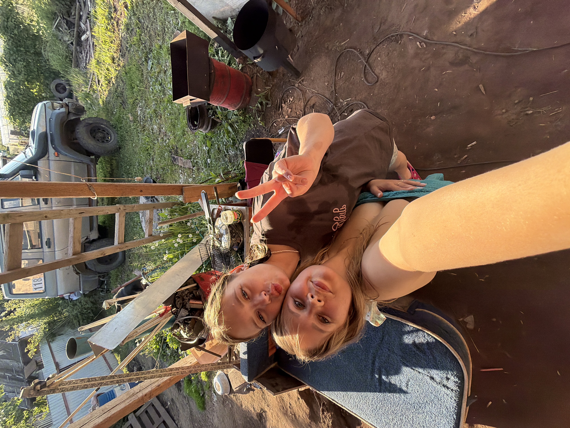 The image depicts two young Caucasian females taking a selfie in an outdoor, semi-rural setting within Chaikovsky, Russia. The subject in the foreground, with long light-brown hair, is positioned with her right arm extended, implied to be holding the camera. She wears a light-colored top, possibly a swimsuit or tank top, revealing her bare shoulder, and displays a subtle smile.
Behind and slightly to her right is a second female subject, with darker brown hair that appears wet. She rests her head against the first subject's, making a "duck face" or puckered lips expression. Her right hand is raised, displaying a peace sign (V-sign) with light-colored nail polish. She wears a dark brown short-sleeved t-shirt with partially visible light-colored text ("Chih" or similar stylized letters) and a simple beaded necklace.
The subjects are positioned on or next to a blue, textured cushion or thin mattress, which rests on an unseen base. The immediate ground beneath is dark brown, dry earth, scattered with small organic debris and pebbles. A dark electrical cable coils on the ground to the right of the foreground subject's arm.
The background reveals a cluttered outdoor area under strong, warm, natural sunlight, suggesting late afternoon or early morning. To the left of the subjects, a collection of haphazardly stacked materials includes wooden planks, metal sheets, a partially visible blue barrel, a green cylindrical bin, and a red container filled with miscellaneous scrap metal. A dark object, possibly a small black dog, is discernible amidst this debris.
In the central background, a drab green/grey SUV, resembling a UAZ Patriot, is parked on a grassy area. It features large off-road tires, a front snorkel air intake, and a white '118' sticker affixed to its rear passenger window. A rustic wooden ladder leans vertically nearby. To the right, a makeshift barbecue setup consists of a rectangular metal brazier resting on a red barrel, which in turn sits on a tire rim. A black, cylindrical metal object, possibly a stovepipe or container, is on the ground nearby. The area is overgrown with grass and white-flowered weeds. Behind the vehicle and extending across the background is a dense green hedge or thick vegetation, with sections of corrugated metal fencing also visible.
