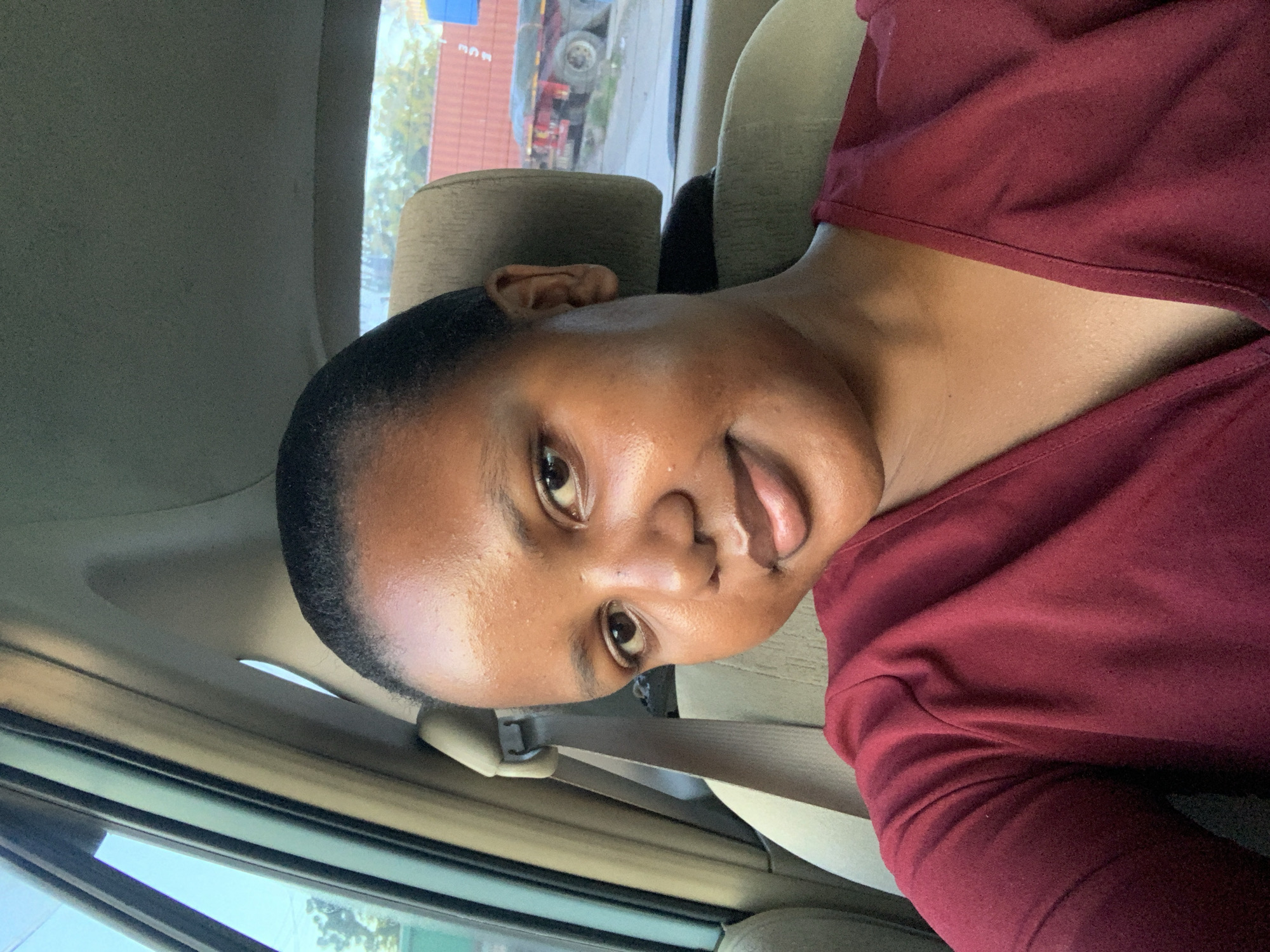 A person with dark skin and short, dark hair is sitting in a car. They are wearing a dark red top. The interior of the car is beige, and a seatbelt is visible. Outside the car window, a truck is partially visible, along with some green vegetation. It is not possible to confirm this is Dar es-Salaam, Tanzania.