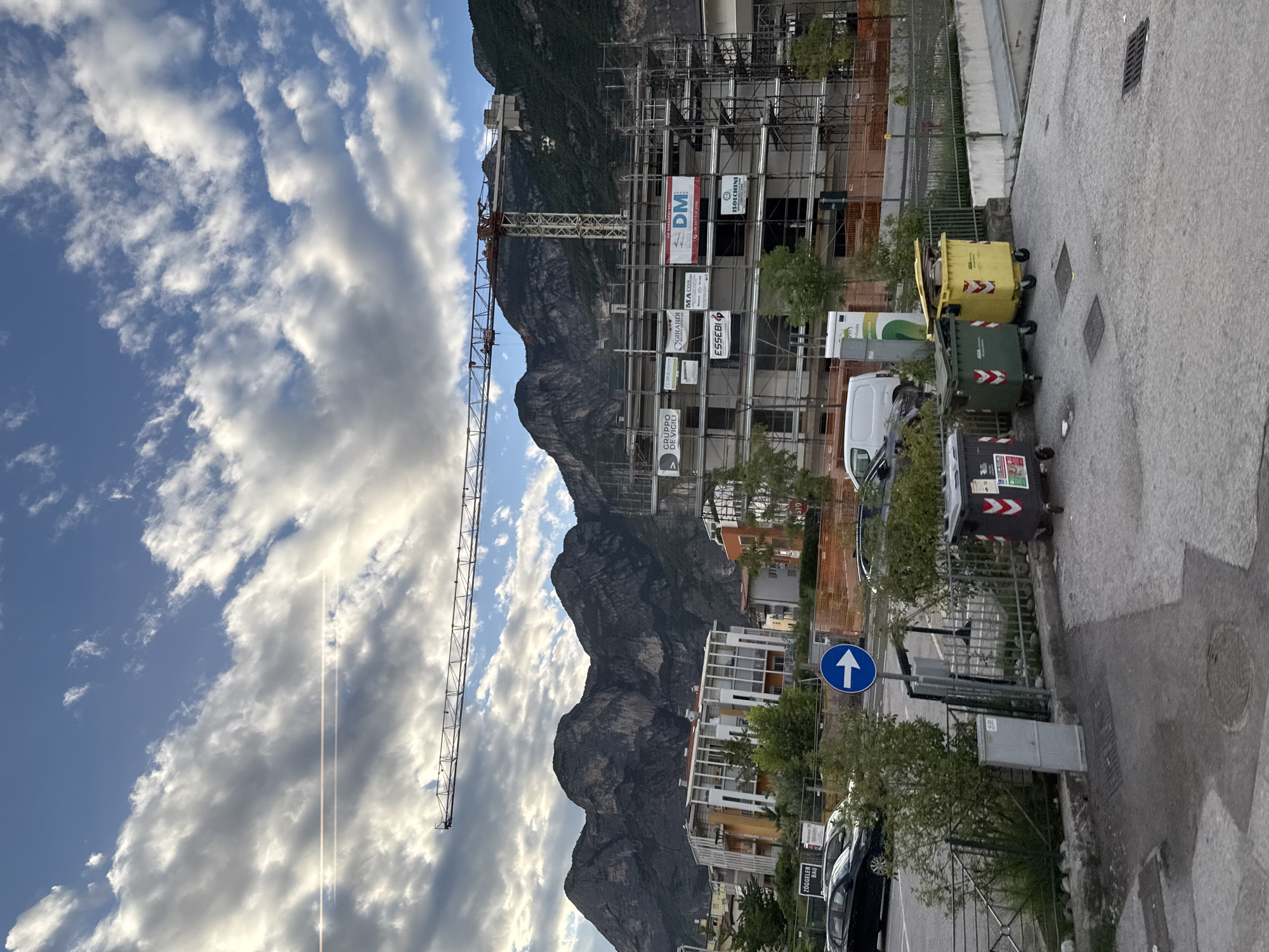 This image depicts a construction site with a large crane prominently featured against a backdrop of a mountain range and a cloudy sky. The scene is oriented vertically. The construction site appears to be in an urban or semi-urban setting, with several buildings under construction, some partially clad in scaffolding and orange safety netting. Signs are visible on the scaffolding, including "DM," "MA CO.S.," "GIRARDI," "ESSEBI," and "GRUPPO DE VIALI."
In the foreground, to the right, are several large waste bins. One is yellow, and two are dark green and grey, respectively, with reflective arrows on their sides. A white van and other vehicles are parked along the road. A blue traffic sign with a white upward-pointing arrow is visible. The overall impression is of an active construction project in a mountainous region.
The time of day appears to be late afternoon or early evening, given the angle of the sun and the quality of light illuminating the clouds. The sky is a mix of blue sky and substantial white and grey clouds, suggesting varied weather. The location is identified as Mezzolombardo, Italy, which is consistent with the mountainous scenery and the presence of Italian text on the signs. No people are clearly visible in the image.