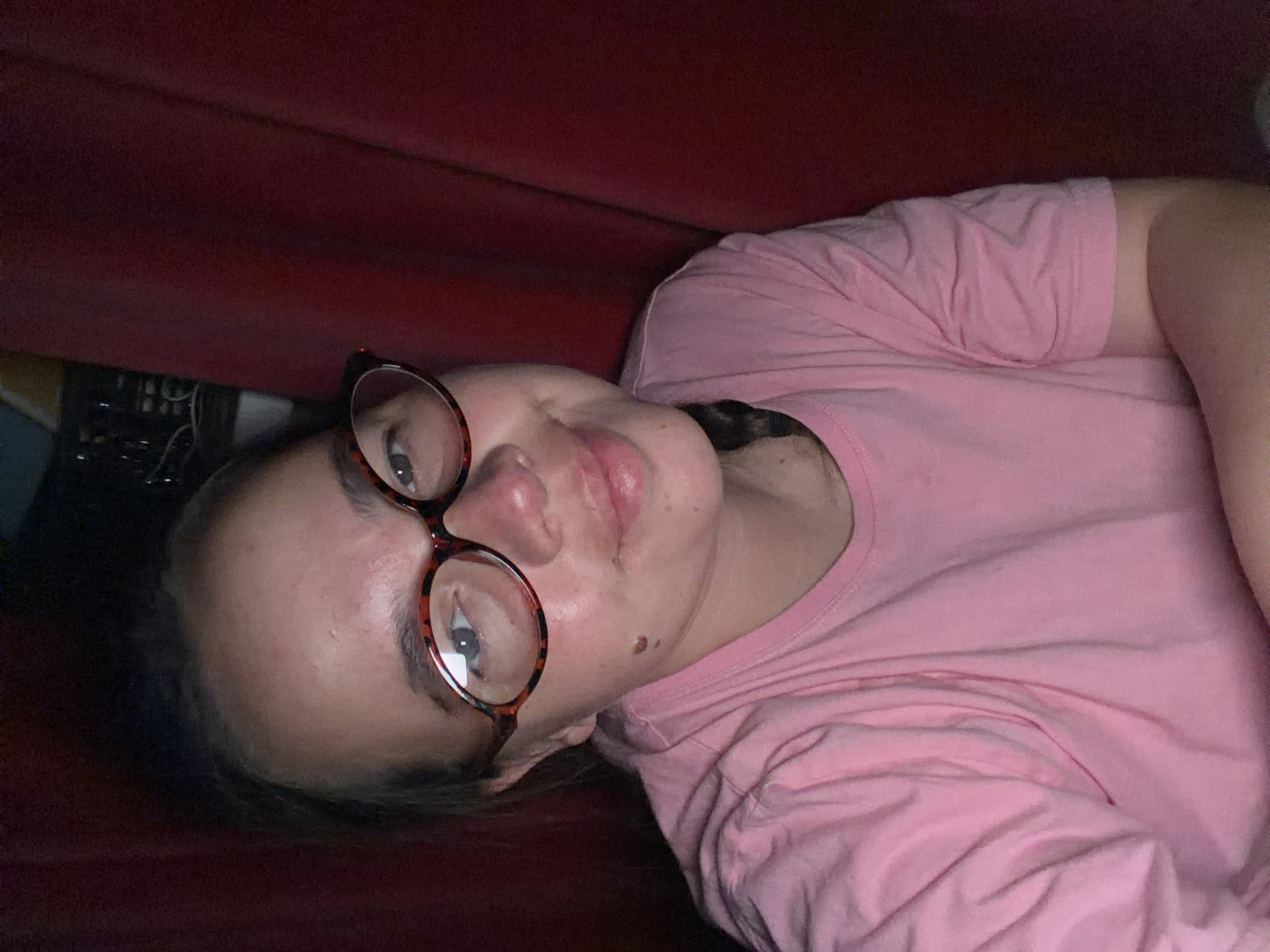 A person wearing eyeglasses and a pink t-shirt lies on a red surface. The setting appears to be indoors, and the environment is dimly lit. It is difficult to identify the precise location in the city of Chaikovsky, Russia without additional information.