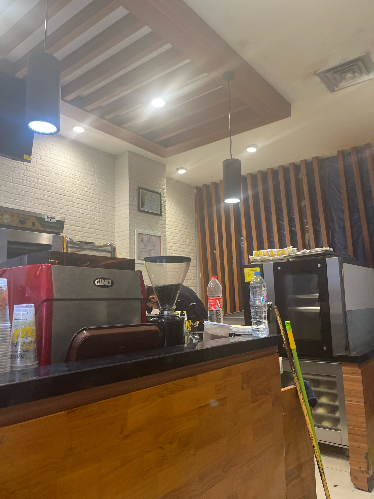 This image depicts the interior of a coffee shop, likely in the city of Sengkang, Indonesia. The scene is dominated by a wooden counter with a dark countertop. On the counter, several items are visible: a red and silver coffee machine with the brand name "GINO" prominently displayed, a coffee grinder filled with beans, stacked disposable cups, a bottle of water, and a Coca-Cola bottle. Behind the counter, a person is partially visible, appearing to be working. In the background, a brick-patterned wall is present on the left, and a wall with vertical wooden slats covered in dark plastic is on the right. Above, a modern ceiling with wooden beams and recessed lighting is observed. There is also a visible air vent in the ceiling. Two brooms are leaning against the counter on the right side of the image. The overall impression is of a functional and somewhat utilitarian coffee shop environment.
