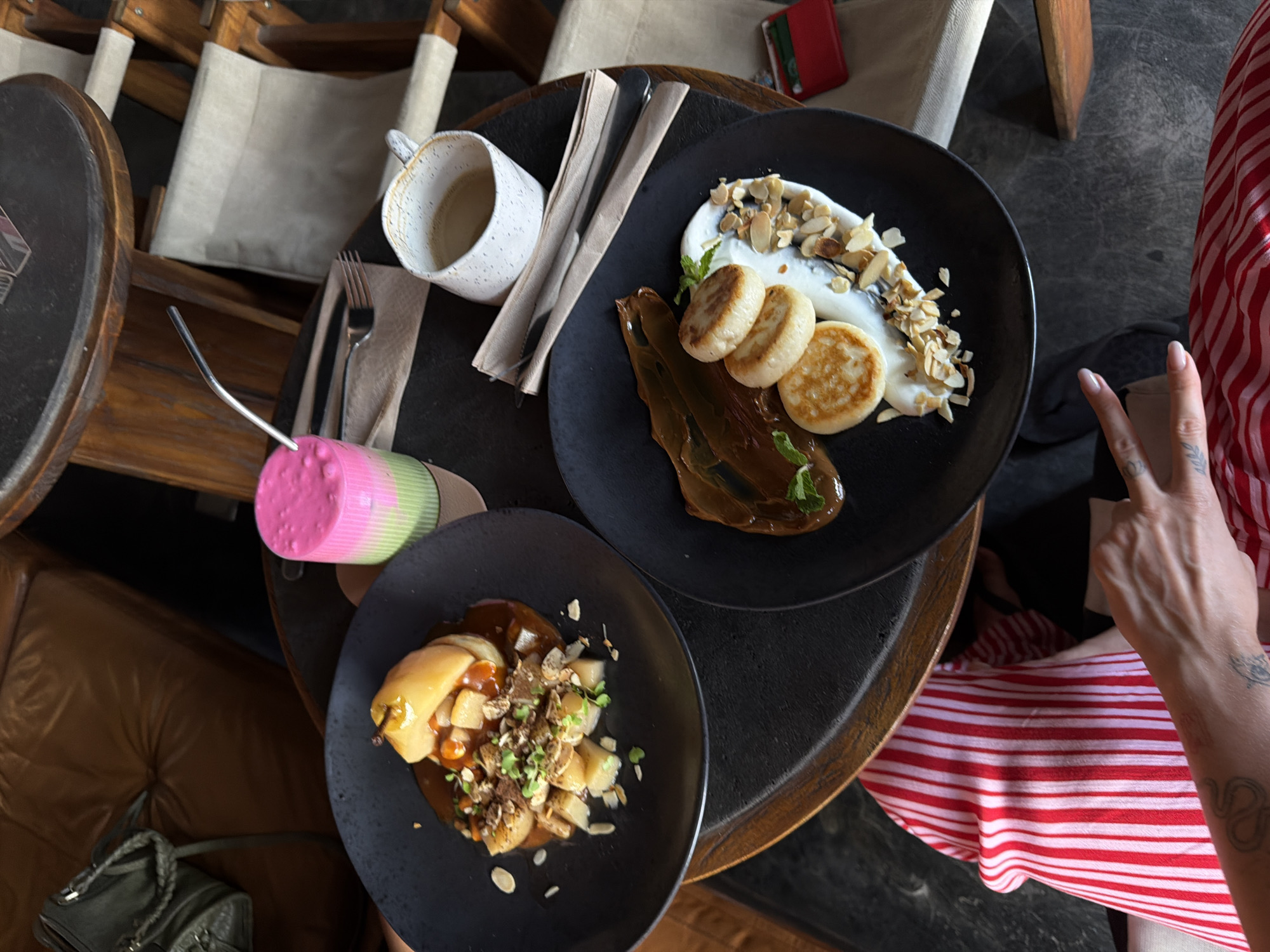This media file shows a top-down view of a table setting in what appears to be a cafe or restaurant in North Kuta, Indonesia. Two plates of food are visible on a dark circular table, alongside a white speckled mug with some liquid in it and cutlery. The first plate, to the right, contains what looks like a dessert with yogurt, small round pastries, and sliced almonds. The second plate, to the left, holds a portion of what seems to be a pear dish with a caramel-like sauce, topped with nuts and microgreens. A brightly colored pink and green straw is also visible. In the background, out-of-focus chairs with light-colored fabric and wooden frames suggest an indoor or covered outdoor seating area. A person's hand with manicured nails and tattoos is visible in the lower right corner, wearing a red and white striped garment. The overall impression is one of a meal being served, possibly breakfast or dessert, in a casual setting. The lighting suggests it might be daytime.