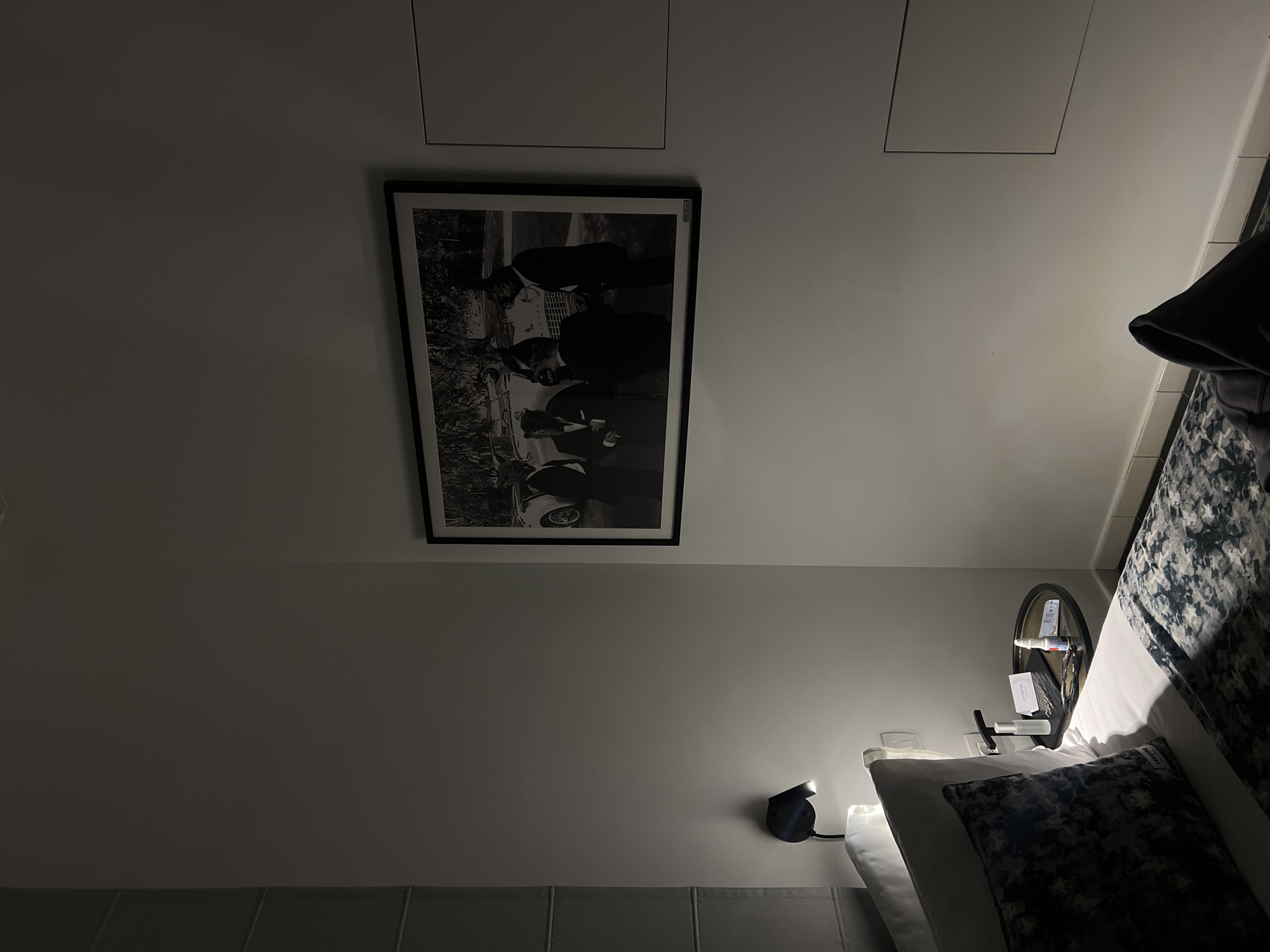 This image depicts a dimly lit bedroom interior, stated to be located in Tel-Aviv, Israel. A white bed with a patterned dark grey and white duvet and pillows occupies the left foreground and midground. A small, round, dark-surfaced nightstand is positioned to the right of the bed. On the nightstand, from left to right, are a small white spray bottle, a black rectangular tray holding small metallic grooming tools (e.g., scissors or clippers) and a folded white card, and a small white bottle with a blue cap (likely nasal spray or eye drops). Above the nightstand, a dark, wall-mounted adjustable lamp provides the primary illumination, casting light downward onto the bedside items.
Centrally on the off-white wall hangs a large, framed black and white photograph. The photograph portrays five figures in dark suits wearing animal heads (specifically tiger or lion heads) gathered around a classic light-colored convertible car. Four figures are clearly visible, with one partially obscured. To the right of the photograph, two rectangular, flush-mounted access panels are visible on the wall. A light grey, vertically pleated blind or curtain structure is present on the far left wall. The bottom of the wall shows a light-colored tiled baseboard. A dark-colored hooded garment is visible in the lower right foreground, resting on the bed or floor. No people are present in the immediate scene; the only human-like figures are within the framed artwork.