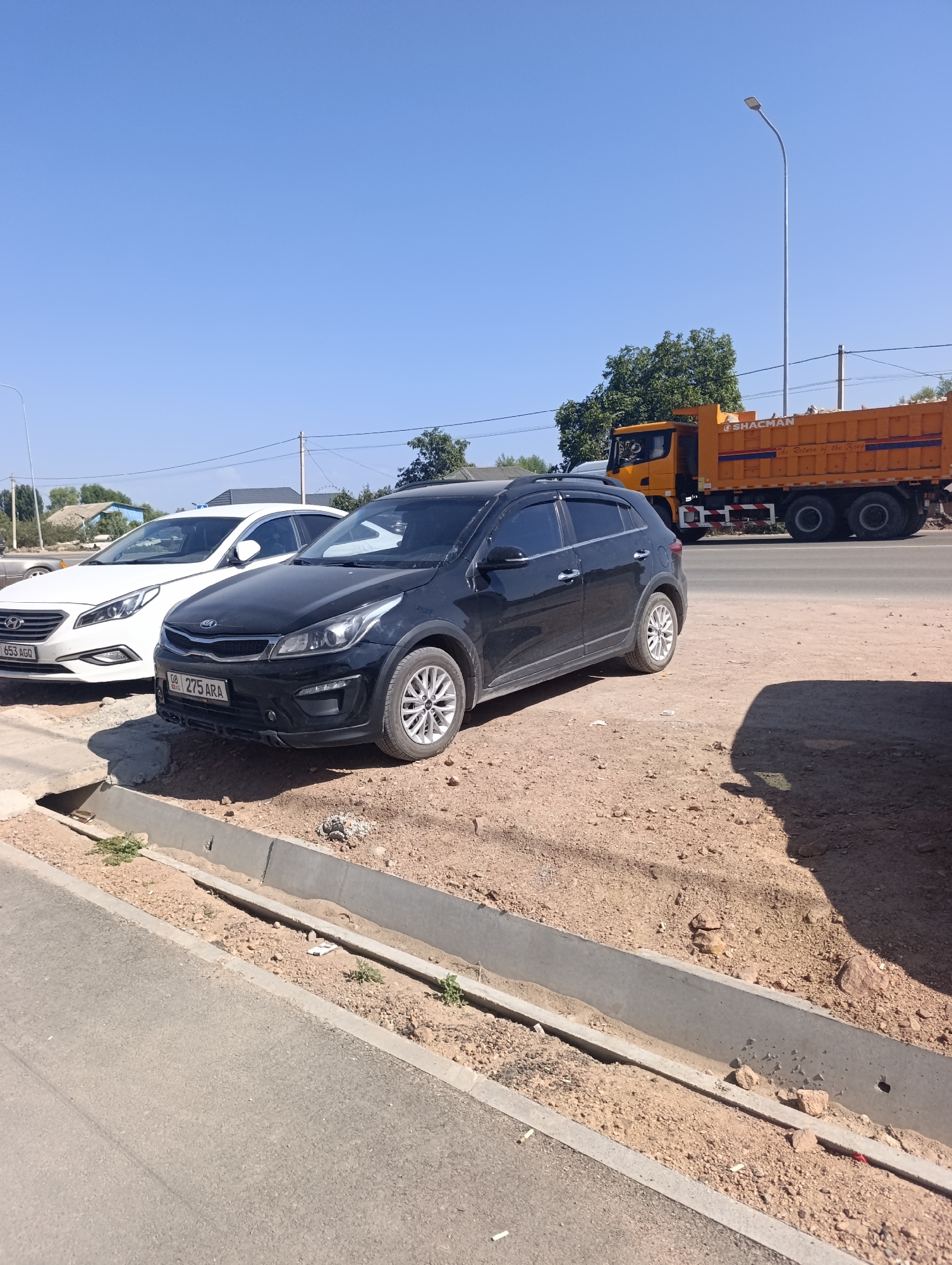 Two parked cars, a black Kia Rio and a white Hyundai Sonata, are visible on a dirt lot next to a paved road. The Kia Rio is in the foreground, with its front end angled towards the viewer. The Hyundai Sonata is parked behind the Kia Rio, to the left. Both cars appear to be in good condition. In the background, a large orange dump truck is driving on the road. The truck has the name "SHACMAN" written on its side. The scene is set against a clear blue sky and a backdrop of trees and buildings. The location is identified as Bosteri, Kyrgyzstan.