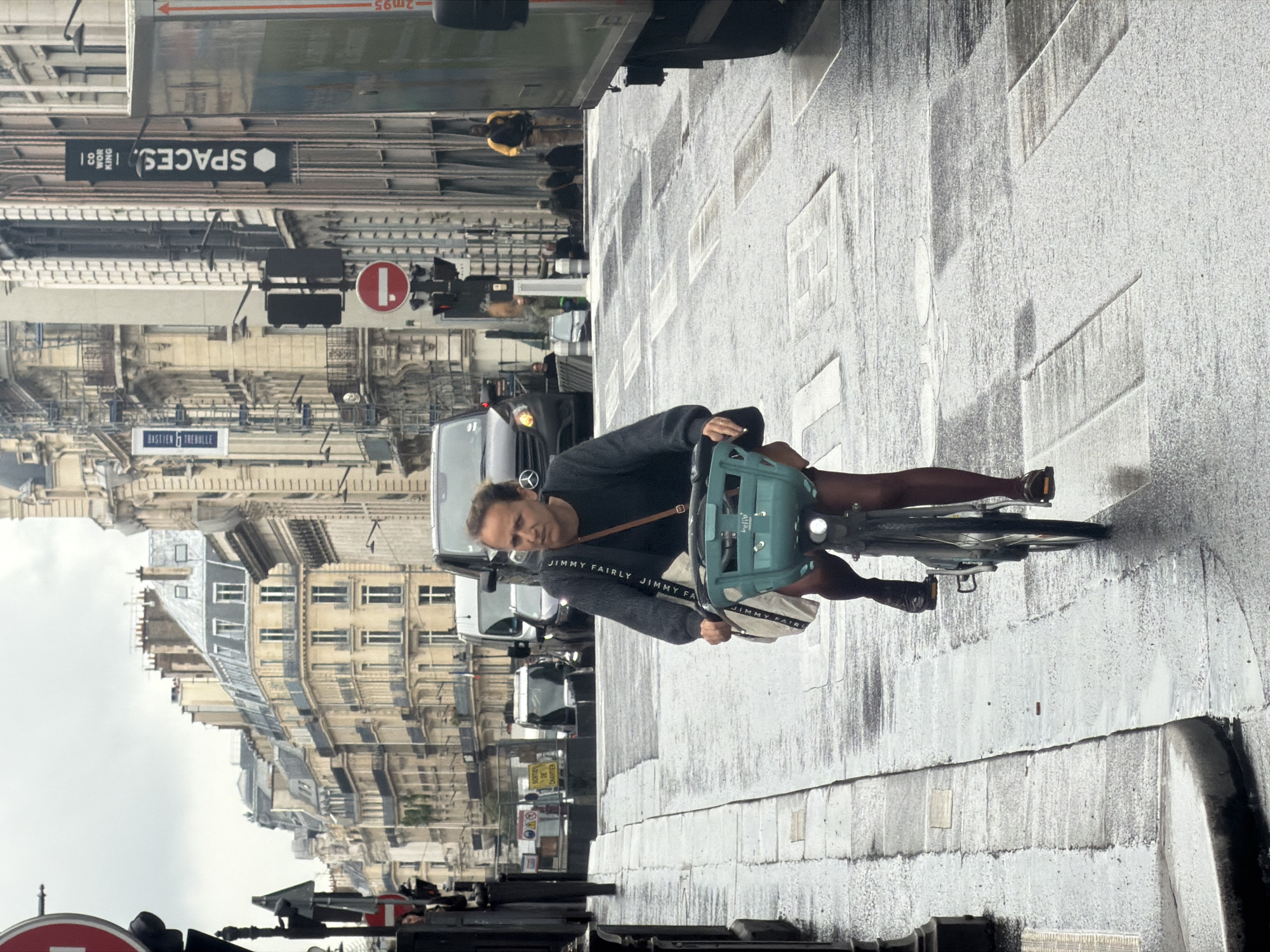A man is riding a bicycle on a wet street in Paris, France. The street is lined with buildings, and there is a "Spaces" office building and various vehicles in the background. The man is wearing a gray sweater, brown shoes and carrying a light teal green bicycle basket and a cream color bag. The street shows wet pavements with rectangular patterns. A road sign with a red circle is on the left.