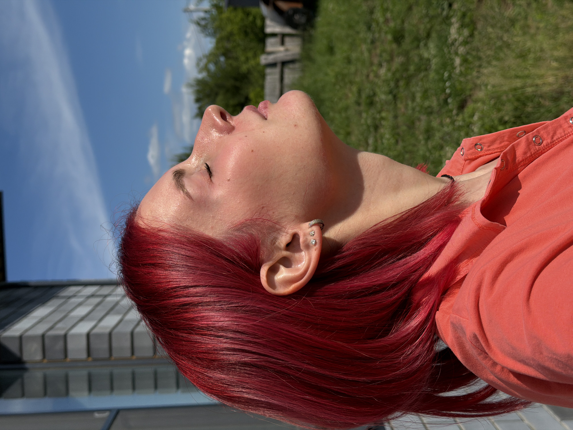 A person with bright, vivid red hair is shown in a close-up profile view, facing upwards with eyes closed, bathed in direct sunlight. Their skin appears flushed and has a slight sheen. The individual wears a coral-colored collared shirt with metallic snap buttons down the placket and a thin, dark choker necklace. Three small earrings are visible on the earlobe: two stud earrings and one small hoop earring.
The background features a clear blue sky with sparse, wispy white clouds. To the left, part of a building with light-colored horizontal siding or brickwork and a dark roofline is visible, along with a reflective window. The ground area is covered in green grass, interspersed with patches of white flora. In the mid-distance, a weathered wooden fence and various green trees and shrubs are present. An indistinct dark object is partially visible on the far right, near the fence line.