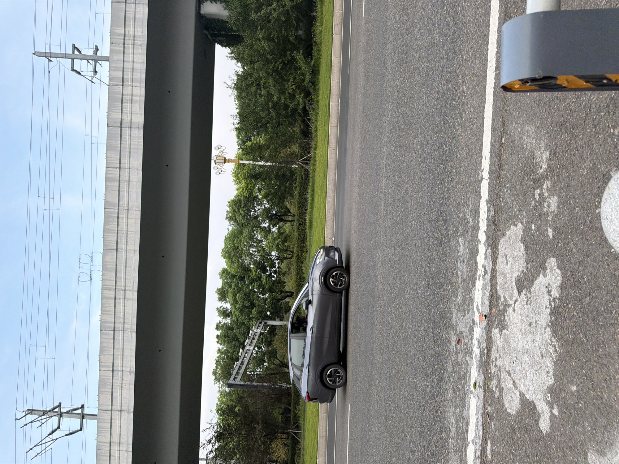 A grey sedan is traveling on a road in Jiangjin, China, beneath an elevated highway. The car is positioned in the right lane. The environment consists of the road surface, a grassy median with trees and foliage, and the concrete structure of the overpass above. Overhead power lines are visible against a cloudy sky. A decorative street lamp stands to the right of the road, and a concrete barrier with yellow and black markings is partially visible in the foreground on the right.