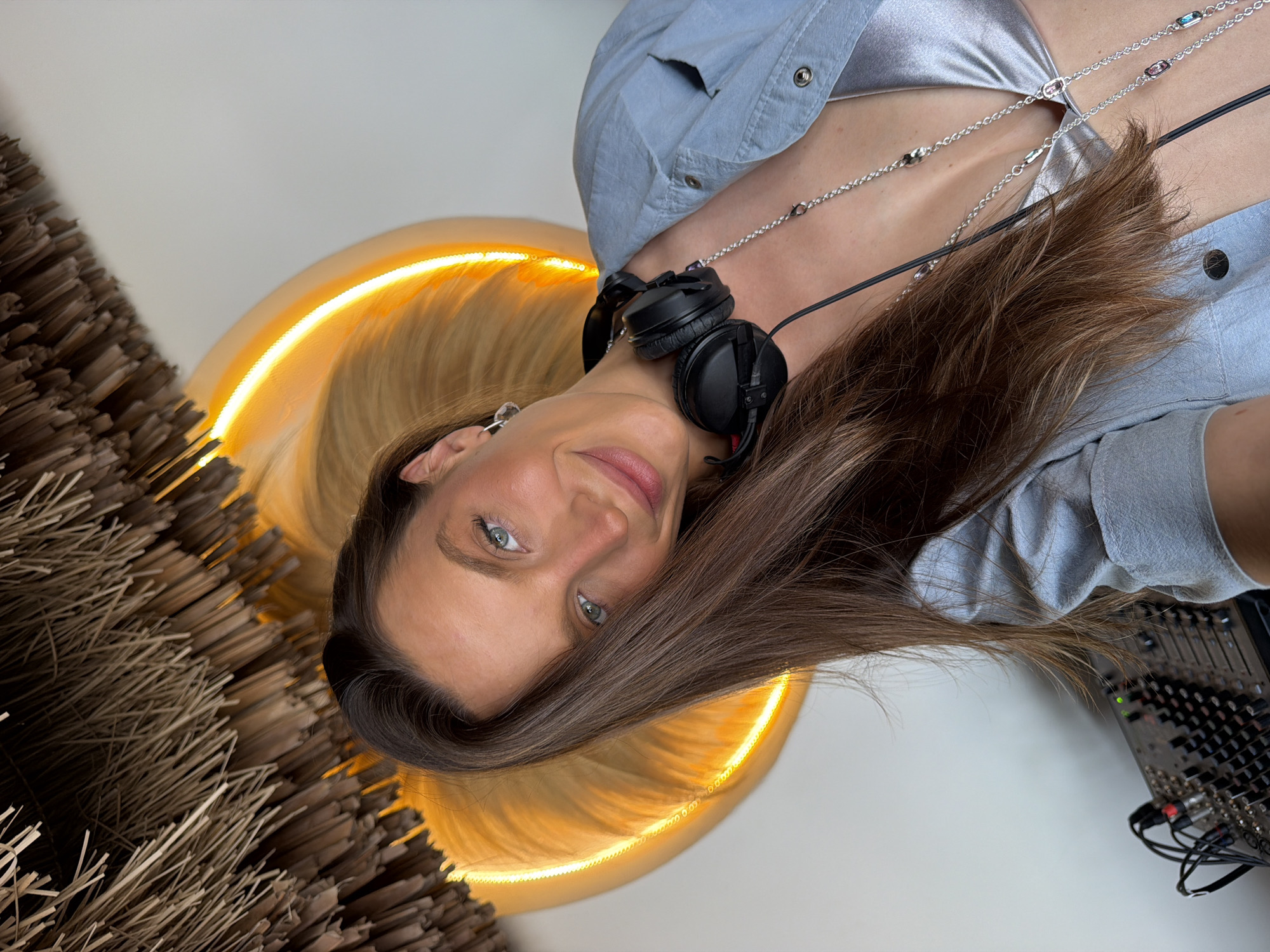In Drobnići, Montenegro, a young woman with long, brown hair and blue eyes smiles at the camera, wearing a light blue jacket over a silver top. She has black headphones around her neck and is adorned with a silver necklace. To her left, a sound mixing board is visible. Behind her, a circular, illuminated structure glows with orange light, and a thatched ceiling is overhead.