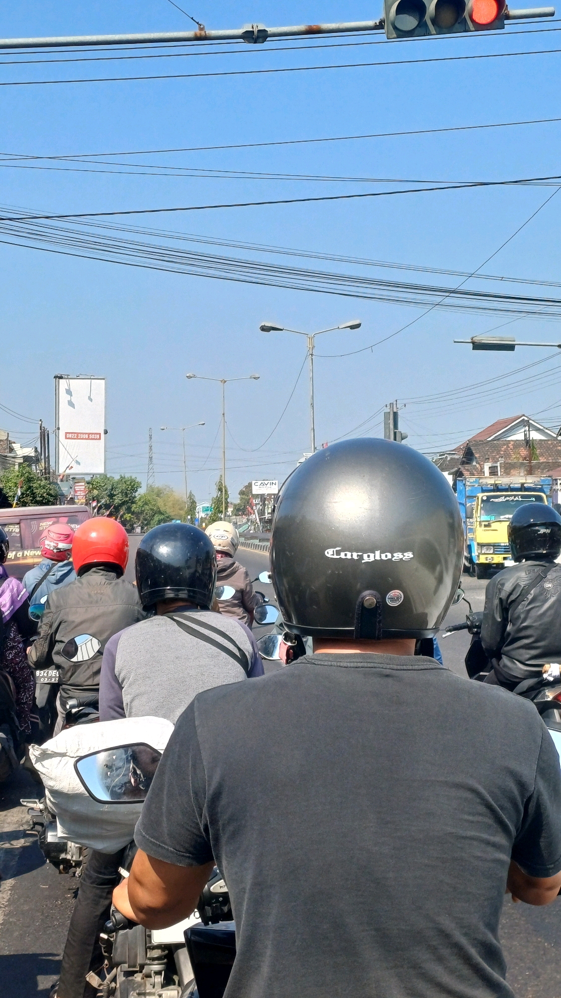 A group of motorcyclists are stopped at a traffic light on a sunny day. The scene is captured from behind a motorcyclist wearing a dark grey helmet with "Cargloss" written on it. The motorcyclist in front of them is wearing a black helmet and a grey shirt. Other motorcyclists are visible further ahead in the frame. In the background, there are traffic lights, street lamps, power lines, buildings, and a yellow truck. The image appears to have been taken in Delanggu, Indonesia, based on the context provided.