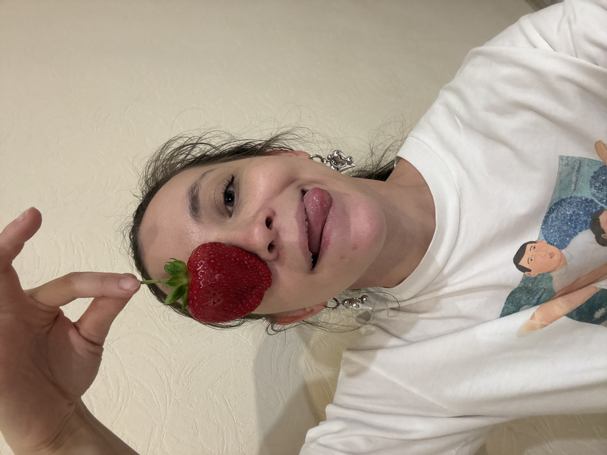 A light-skinned woman with brown hair, wearing a white t-shirt with a print of people on it, poses for a selfie. She is holding a large strawberry in front of her left eye. Her tongue is sticking out. She wears silver earrings. The background is a light-colored wall. There are no visible indications of the city of Kazan, Russia.