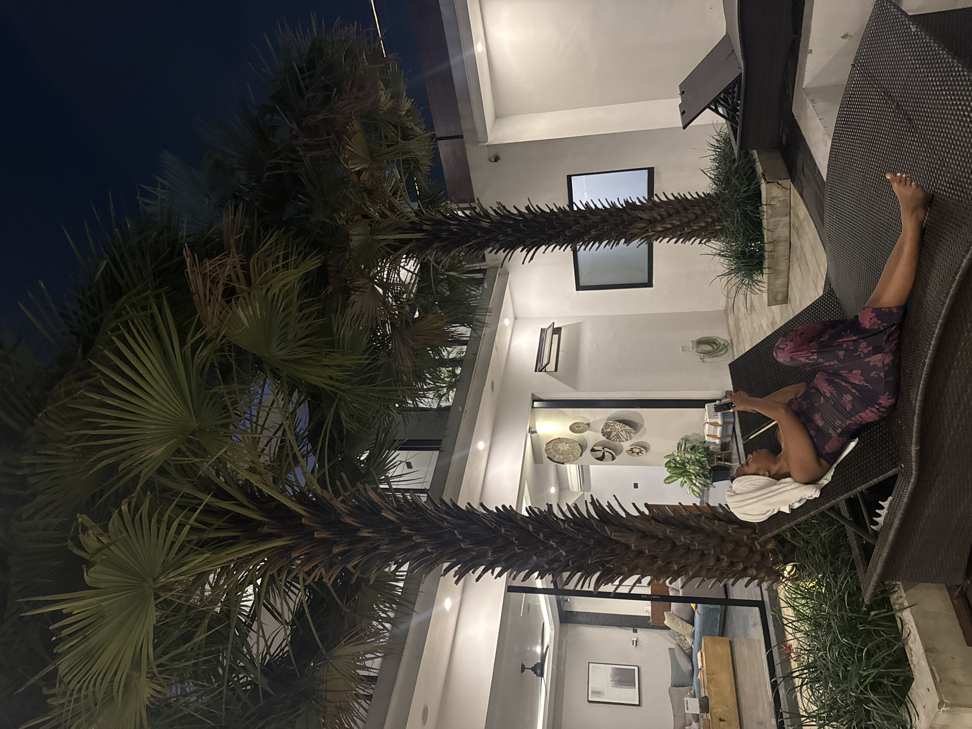 A person lies on a dark brown woven chaise lounge, looking at their phone. They are wearing a dark purple dress with a floral pattern and have a white towel wrapped around their head. The chaise lounge is situated on a paved patio area, surrounded by lush green plants. In the background, a modern building with white walls and large windows is visible. Two tall palm trees with large fronds dominate the foreground, their branches reaching towards the dark night sky. The scene appears to be an outdoor patio at night, illuminated by artificial lights from the building, creating a serene and relaxed atmosphere. The overall setting suggests a vacation or resort location. There is no visible text in the image.
