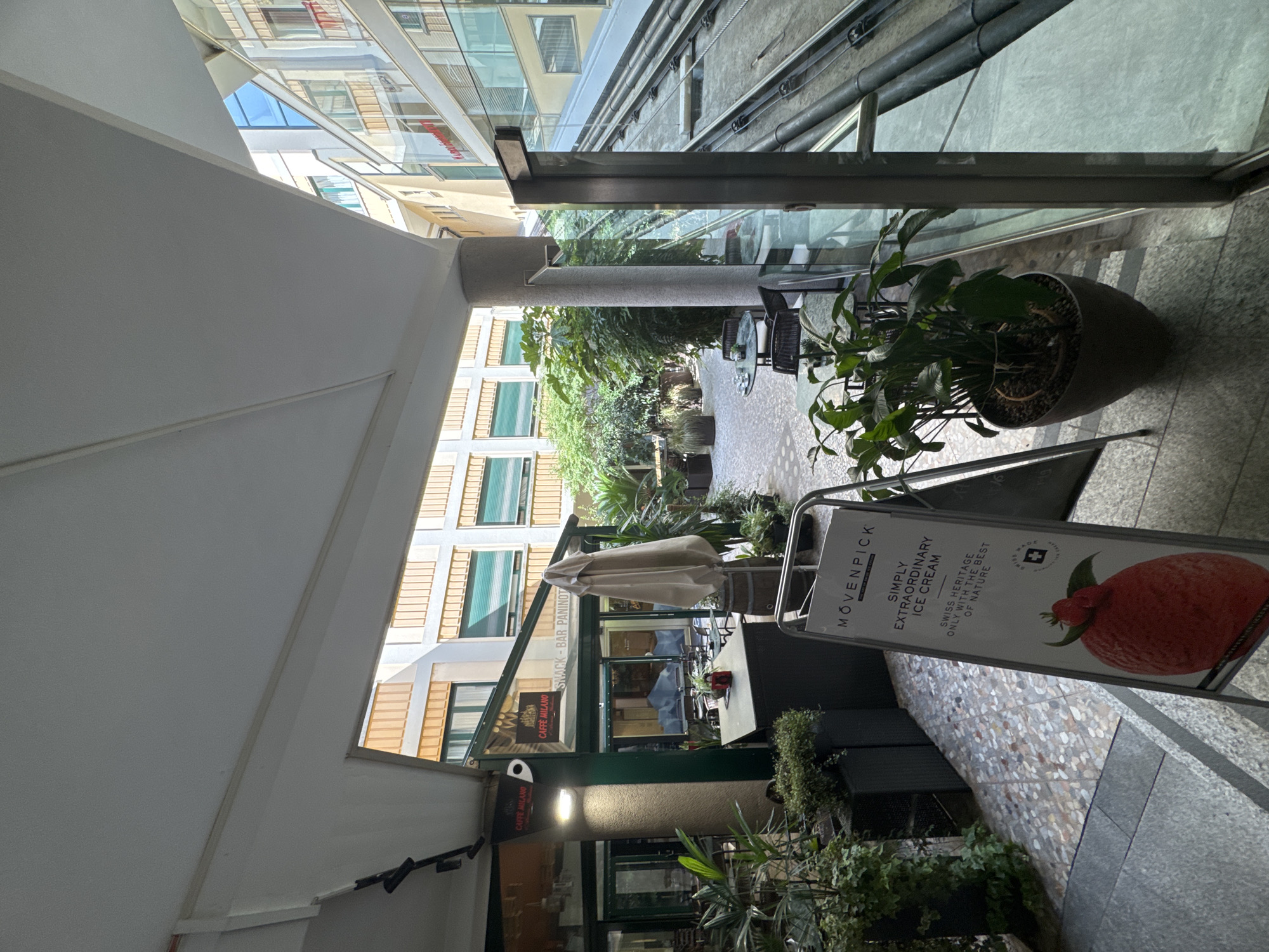 This is an image of the Movenpick Hotel in Lugano, Switzerland, specifically an outdoor seating area. A sign for "Movenpick Simply Extraordinary Ice Cream" is in the foreground, featuring a large strawberry image and text about Swiss heritage. To the left of the sign, a cafe with a green awning and "Caffè Milano" branding is visible, with tables and chairs set up outside. Further back, the hotel's courtyard opens up to reveal architectural elements of surrounding buildings, with windows and balconies. Lush green plants are strategically placed throughout the scene in pots, adding to the ambiance of the outdoor space. The flooring is a mosaic of small, irregularly shaped tiles. The overall impression is a serene and inviting outdoor area within a hotel setting.