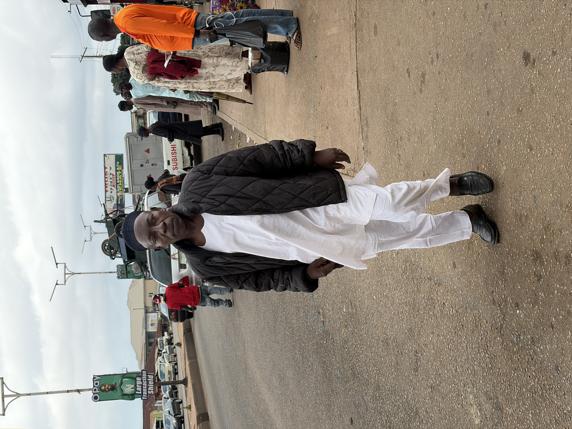 The image shows a man walking on a paved road in Gura Top, Nigeria. He is wearing a black quilted jacket over a white traditional outfit and black loafers. He is looking towards the camera. In the background, there are other people standing on a sidewalk, as well as several vehicles, including cars and a white truck. There are also streetlights and billboards visible. The sky is overcast. The scene appears to be a busy urban environment.
