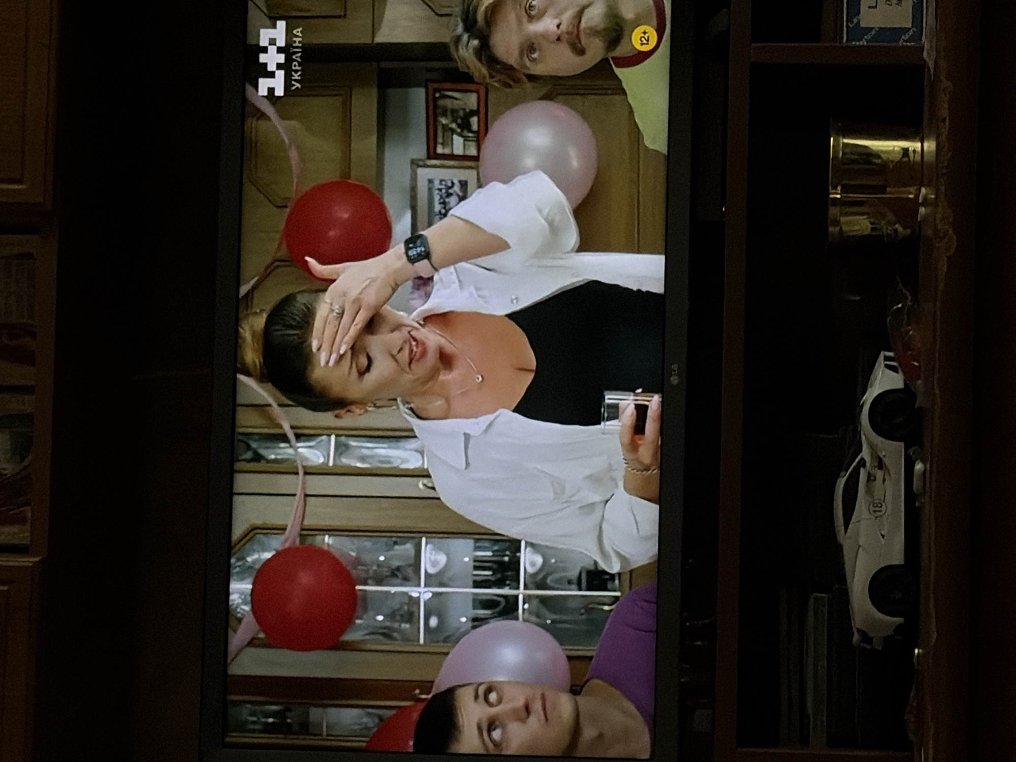 A television screen displays a scene from a show. In the foreground, on a shelf beneath the television, rests a white toy sports car and a gold-colored cylinder. On the television screen, three people are visible. On the left, a young man with short dark hair and light-colored eyes looks up and to his left, wearing a purple t-shirt. In the center, a woman with dark hair pulled back, wearing a white open-collared shirt over a dark V-neck top, is seen from the chest up. She is touching her forehead with her right hand, her eyes are closed, and she holds a clear glass with a dark liquid in her left hand. A silver necklace with a pendant is visible around her neck, and she wears a ring on her right ring finger. A watch with a dark band and digital display is on her left wrist. To the right of the woman, a man with light brown hair and a mustache is visible from the chest up, wearing a yellow t-shirt with a red collar. He looks to his right, with a slightly furrowed brow. Behind the people are several balloons, two red and one pink, and a light-colored wall with framed pictures and glass-paneled doors. A graphic in the upper right corner of the screen displays "1+1 УКРАЇНА" and a "12+" age rating icon is visible on the lower right of the screen.