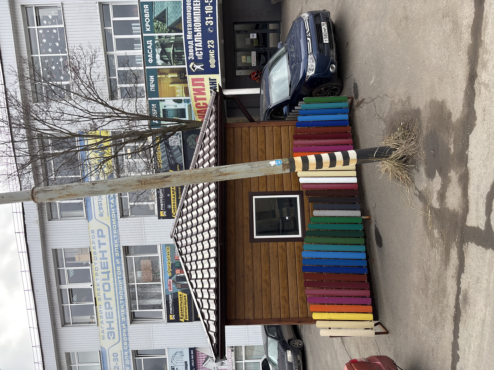An outdoor daytime shot in Izhevsk, Russia, captures a commercial street scene featuring a multi-story building, a small display structure, a parked car, and samples of building materials.
The main building is clad in light grey siding with numerous windows. Its facade is covered with large banners advertising various products and services. Visible text includes "МАГАЗИН ЭЛЕКТРОТОВАРОВ ЭНЕРГОЦЕНТР" (Electrical Goods Store "ENERGOtsentr") with phone number 8(951)2..., and a description "ДЛЯ СБОРКИ ЭЛЕКТРОЩИТОВ И ЭЛЕКТРОМОНТАЖ" (for assembling electrical panels and electrical installation). Other prominent signs advertise "Завод Металлокровли «СТАЛЬКОМПЛЕКТ»" (Metal Roofing Factory "STALKOMPLEKT") with office number "офис 23" and phone number 31-10-5, along with sections for "КРОВЛЯ" (ROOFING), "ФАСАД" (FACADE), and "ПЕЧИ" (OVENS). Another advertisement is for "РУФТРЕЙДИНГ KROVENT" (ROOFTRADING KROVENT) with phone +7 (3412) 47-46-16, mentioning "Кровельная вентиляция" (Roof ventilation) and "Снегозадержатели безопасность" (Snow retainers safety). One window on the building is adorned with white heart and snowflake decals.
In front of the building, a small, brown wooden structure with a dark tiled roof and a single window serves as an outdoor display. Adjacent to this, a vibrant array of colorful metal sheets, likely samples of roofing or siding, are stacked vertically. These samples showcase various hues including red, blue, green, yellow, white, and a distinctive black and white striped section.
A dark blue sedan with the Russian license plate "T580 MP 18" (indicating the Udmurtia region, where Izhevsk is the capital) is parked near the display area. The ground is paved asphalt, showing some cracks and patches of dry grass. A utility pole stands prominently in the mid-ground, partially obscuring the view, and features some faded, unreadable text. Bare tree branches extend across the upper portion of the image, suggesting a dormant season. The sky is overcast, indicating a cloudy daytime setting without direct sunlight.