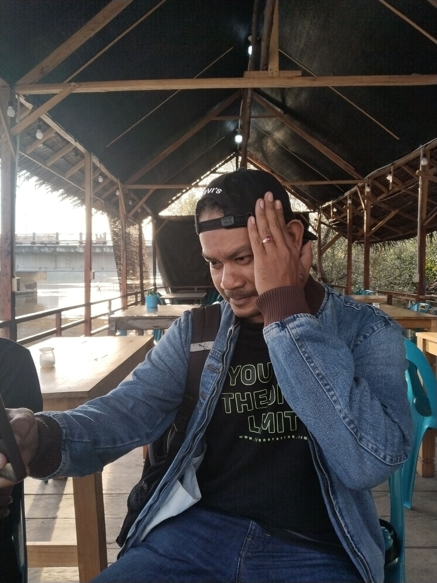 A man is seated at an outdoor cafe table, partially obscured by his hand pressed against his forehead. He wears a blue denim jacket over a black t-shirt with white text that reads "YOU THE LIMIT". A black baseball cap with a red logo adorns his head, and a red ring is visible on his right hand. The cafe appears to be situated near a body of water, with a bridge and a cityscape visible in the background. The lighting suggests it is daytime, possibly late afternoon, with sunlight casting long shadows. The architecture of the cafe features a thatched roof supported by wooden beams, typical of a tropical setting. The specific location is a cafe on the waterfront in Lhokseumawe, Aceh, Indonesia.
