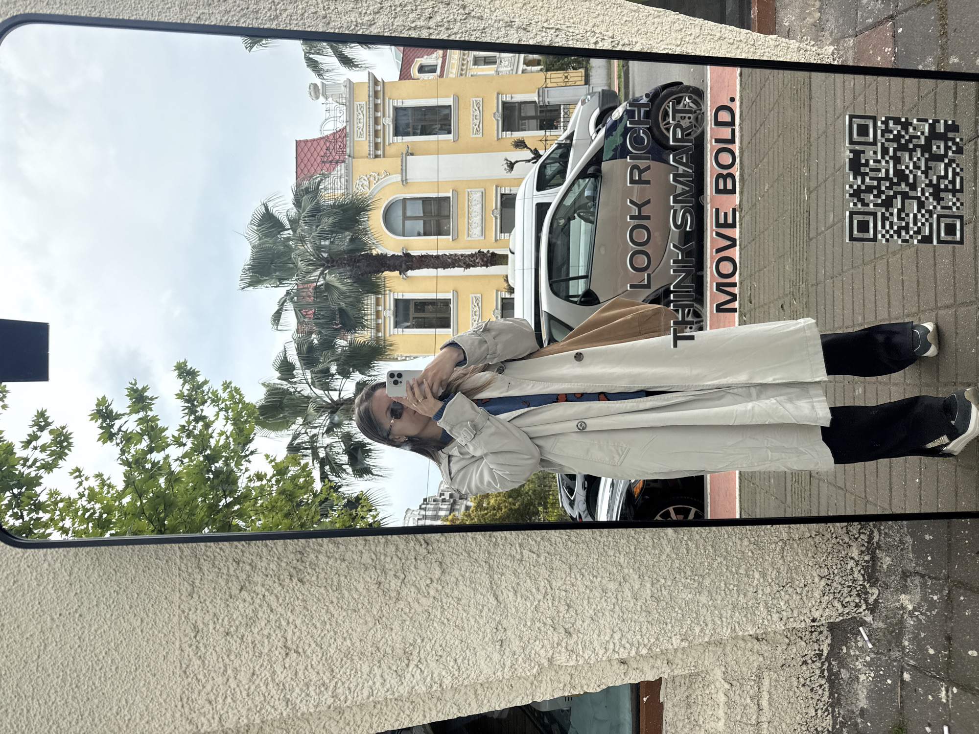 The image displays a large, rectangular outdoor mirror or digital screen, which acts as a reflective surface for the surrounding urban environment. Centrally positioned within the reflection is a young woman, partially obscured by the smartphone she holds up to take a selfie. She is dressed in a light-colored trench coat over a patterned blue top, dark trousers, and sneakers, with sunglasses covering her eyes.
The reflection reveals a lively street scene, characteristic of Batumi, Georgia. In the background, a distinctive yellow building with intricate architectural details and arched windows dominates the view, partially framed by lush green palm trees. Several vehicles, including a white van and a car with a camouflage pattern, are parked along a sidewalk or street. The sky reflected overhead appears bright but overcast.
Prominently featured on the reflected ground surface is an advertisement or public art, displaying bold text that reads: "LOOK RICH", "THINK SMART.", and "MOVE BOLD.". Adjacent to this text is a clear QR code. The visible ground within the reflection is paved with light-colored tiles. Outside the mirror's frame, a textured, light-colored wall surface and dark ground are visible. The scene captures a casual daytime moment, likely during the morning or afternoon, in an outdoor setting.