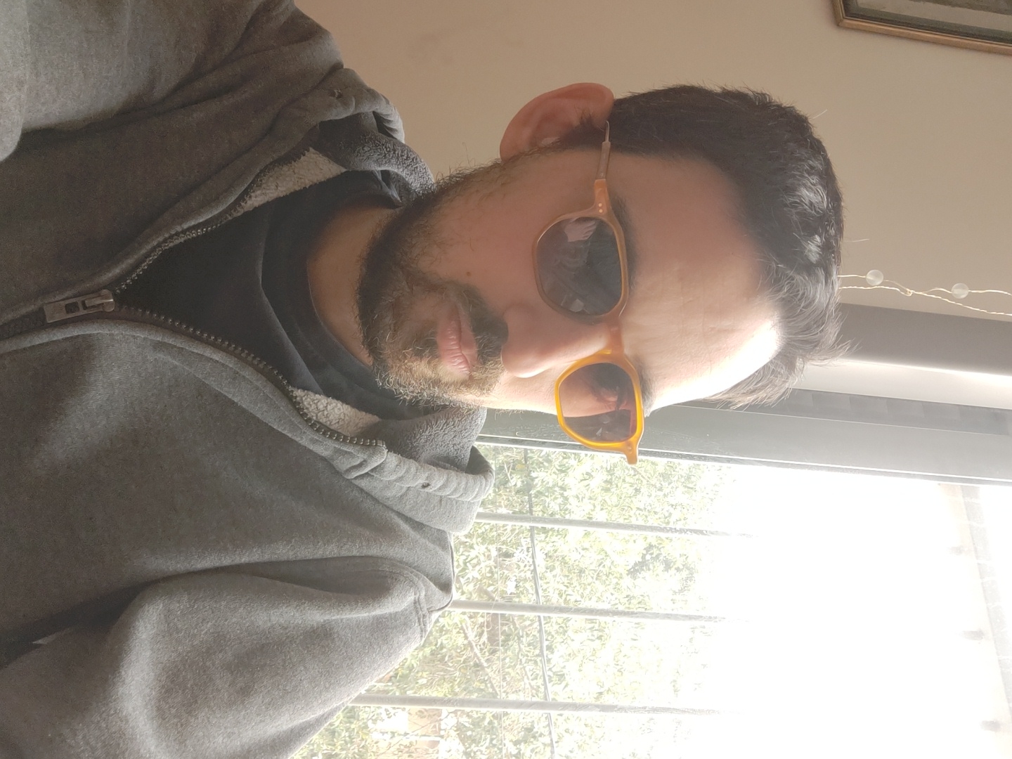 A selfie-style image shows a man with a beard wearing yellow-framed sunglasses. He is wearing a gray hooded sweatshirt and a black t-shirt underneath. A window is visible behind him, and a glimpse of trees is seen through the window. The location is in Ramat Gan, Israel.